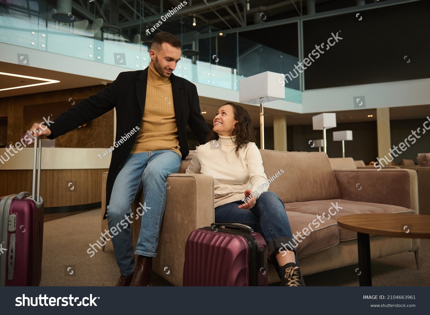 Woman and man in casual clothes with suitcases sitting on armchair in the international airport terminal lounge, looking at each other, discussing forthcoming flight, waiting for airplane departure #2104663961