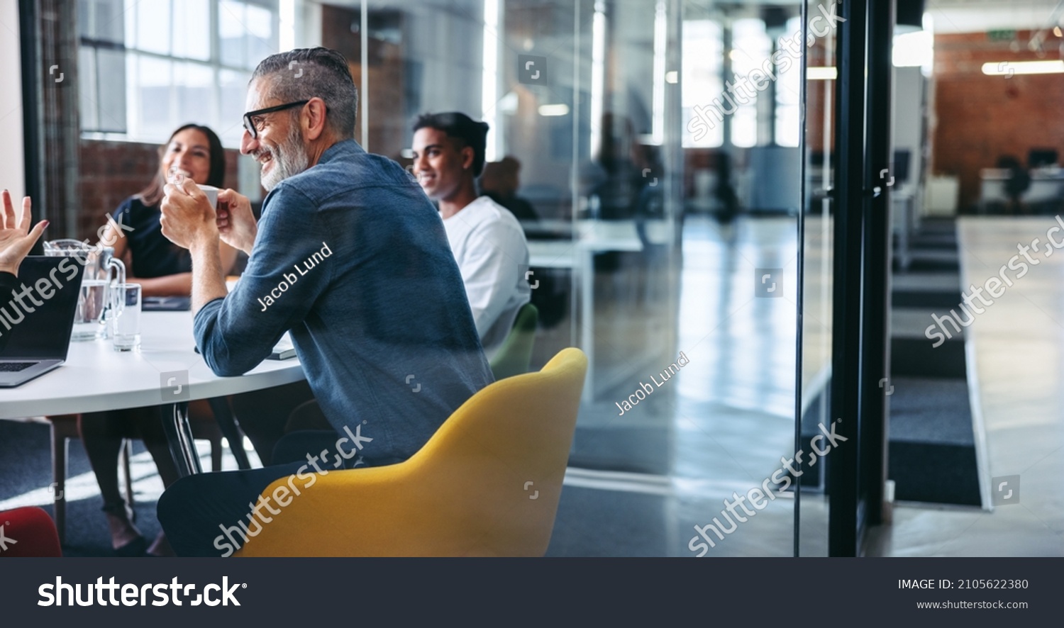 Cheerful mature businessman attending a meeting with his colleagues in an office. Experienced businessman smiling while sitting with his team in a meeting room. Creative businesspeople working together #2105622380