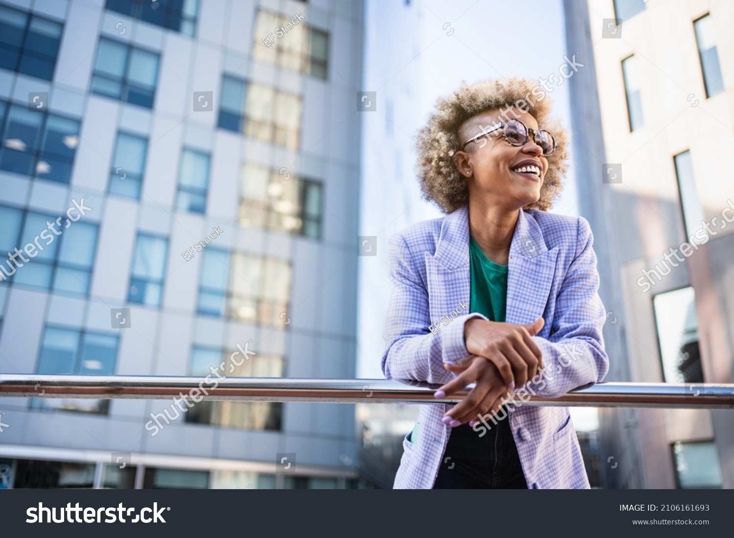 Young African American business woman in trendy outfit and eyeglasses standing on corporate urban district. Inspired creative female professional in the city #2106161693