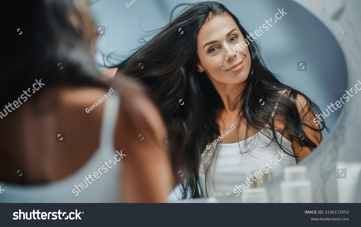 Beautiful Early Middle Aged Woman Looks into Bathroom Mirror Touches Her Lush Black Hair, Admires Her Looks. Concept for Happiness, Wellbeing, Natural Beauty, Organic Skin Care Products #2106172052