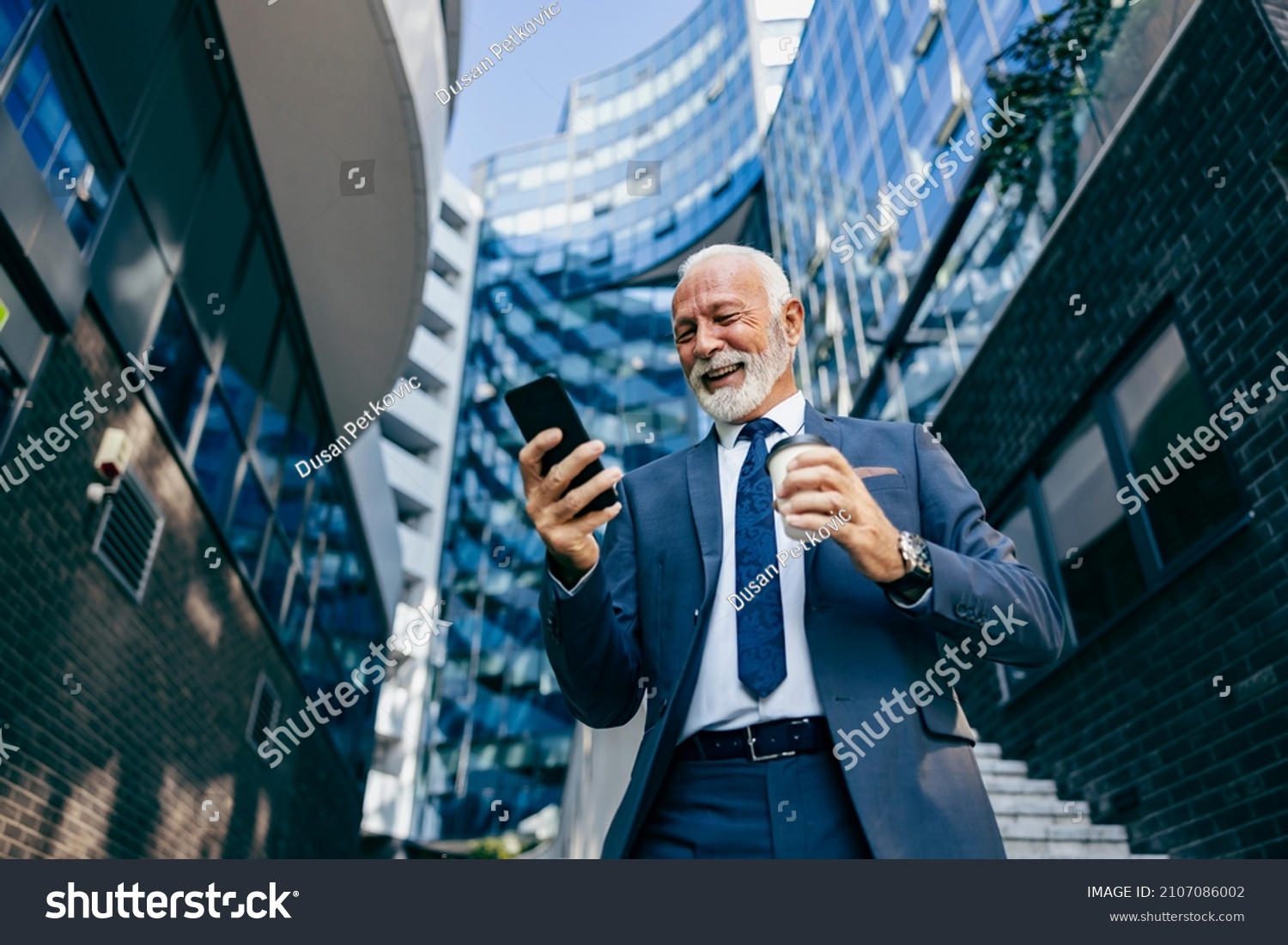 An old executive reading message on the phone at the business center. Low angle view of a happy senior businessman standing at the business center and reading a message on the phone. #2107086002