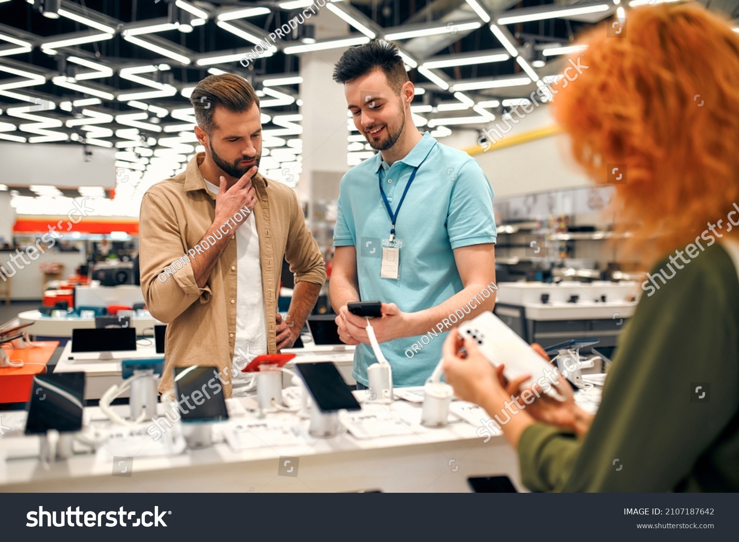 Sales consultant advises man who chooses new smartphone in store of household appliances, electronics and gadgets. Professional consultation in store. Sale Day. #2107187642