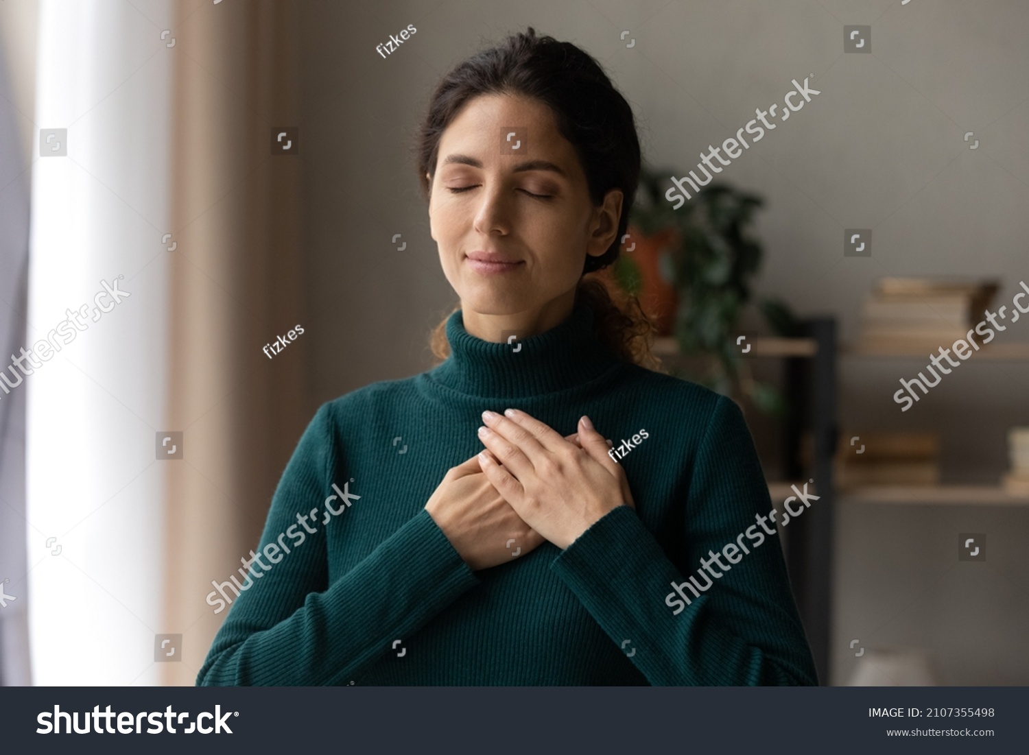 Head shot peaceful young attractive woman holding folded hands on chest, praying waiting for miracle, feeling thankful indoors. Sincere happy female volunteer showing kindness or expressing gratitude. #2107355498