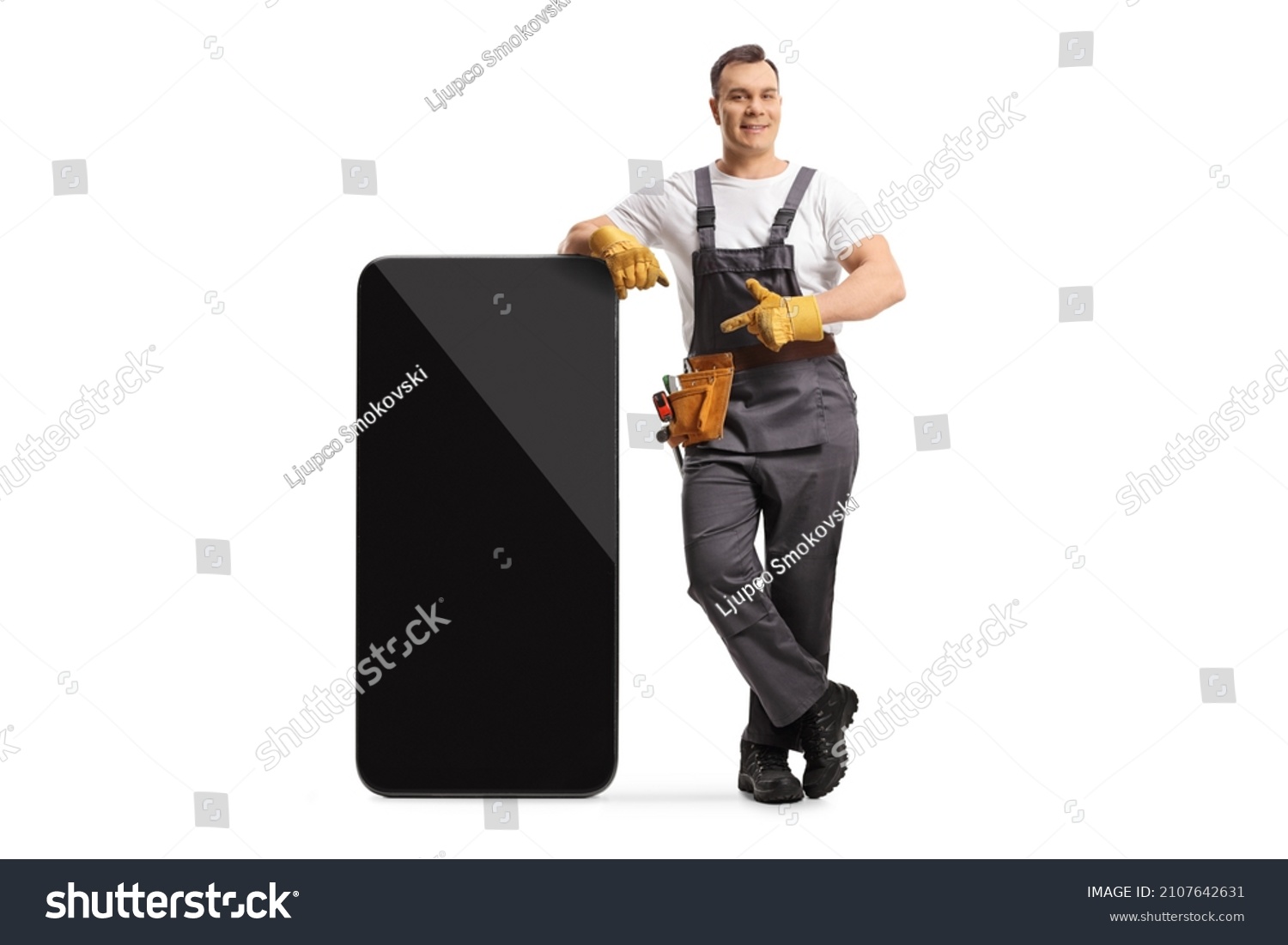 Full length portrait of a repairman in a uniform leaning on a big mobile phone and pointing at the screen isolated on white background #2107642631