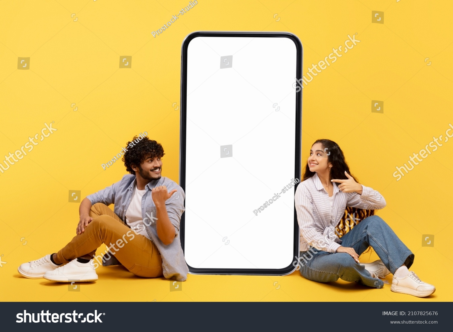 Happy indian couple sitting near big smartphone with blank white screen, demonstrating copy space for app or ad design, posing over yellow background, mockup banner #2107825676