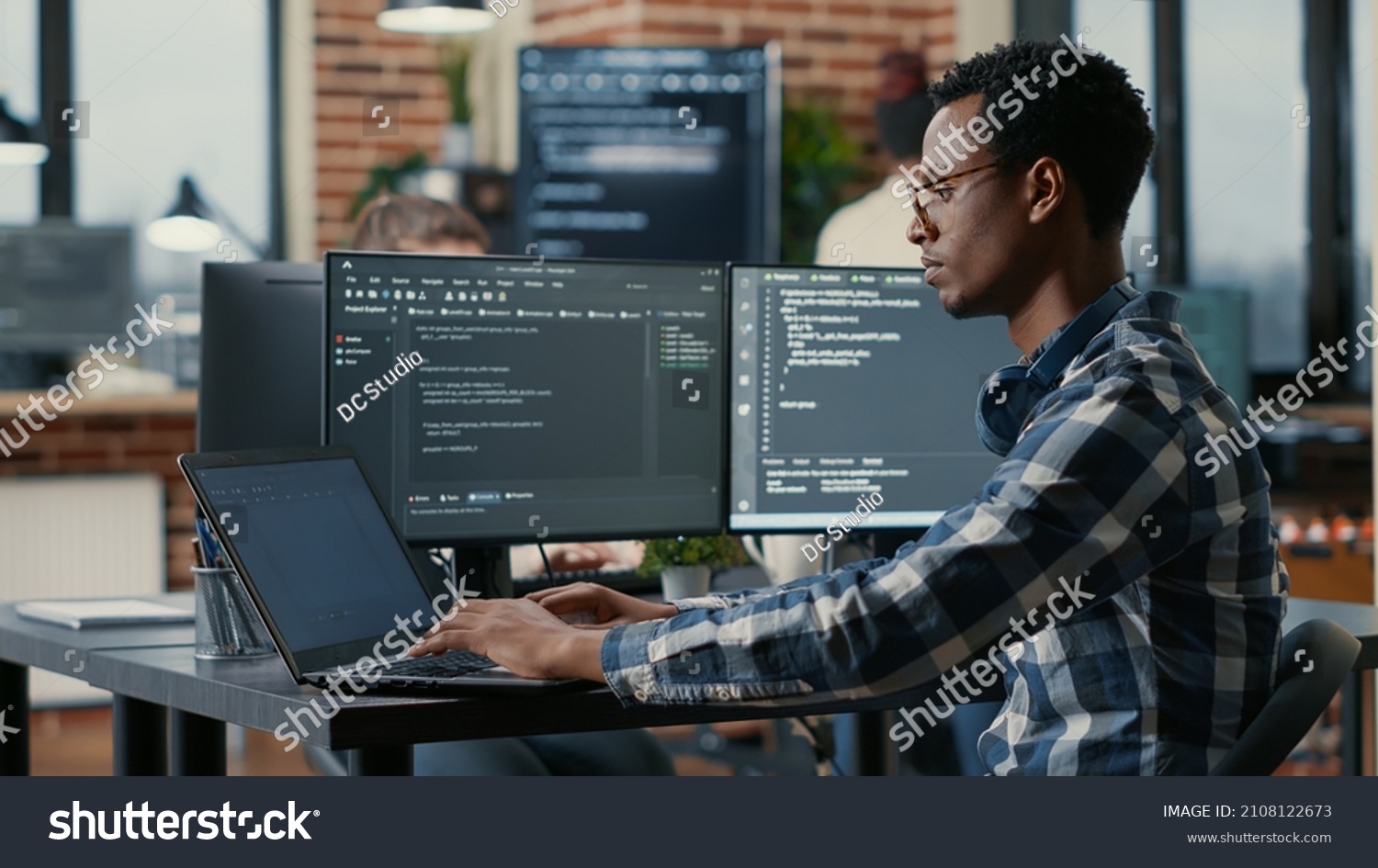 Portrait of african american developer using laptop to write code sitting at desk with multiple screens parsing algorithm in software agency. Coder working on user interface using portable computer. #2108122673