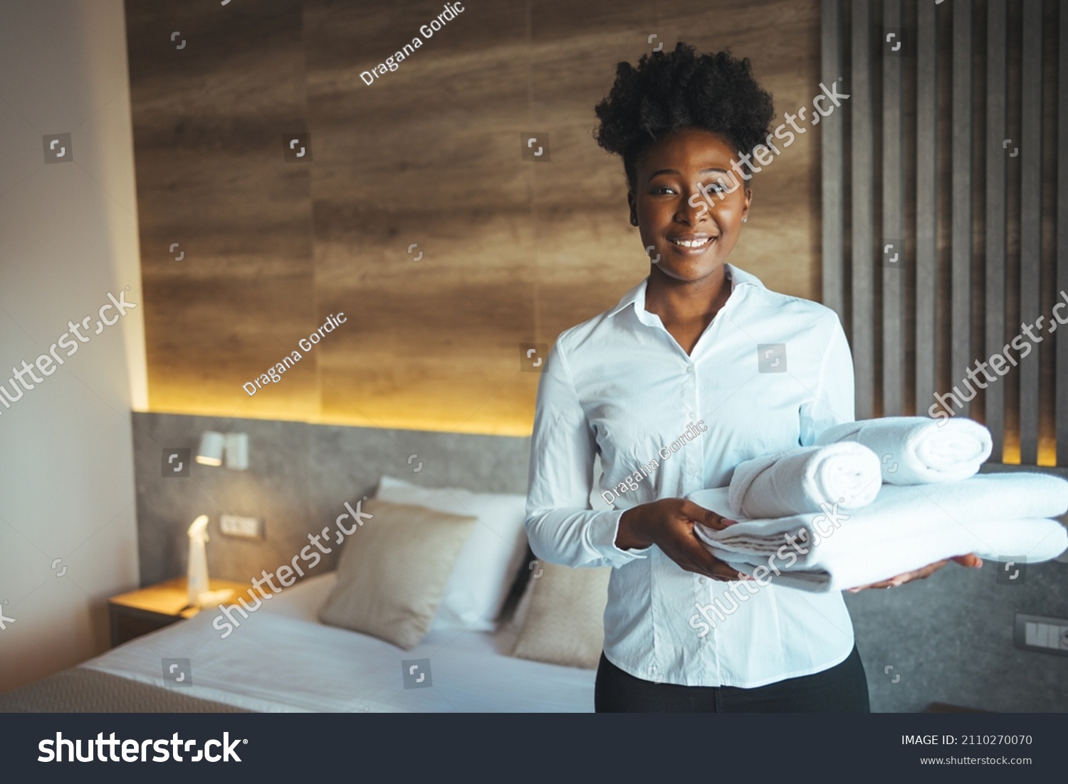 Hotel staff setting up pillow on bed. African housekeeper looking at the camera holding a towel a hotel room. Clean towels during housekeeping in a hotel room #2110270070