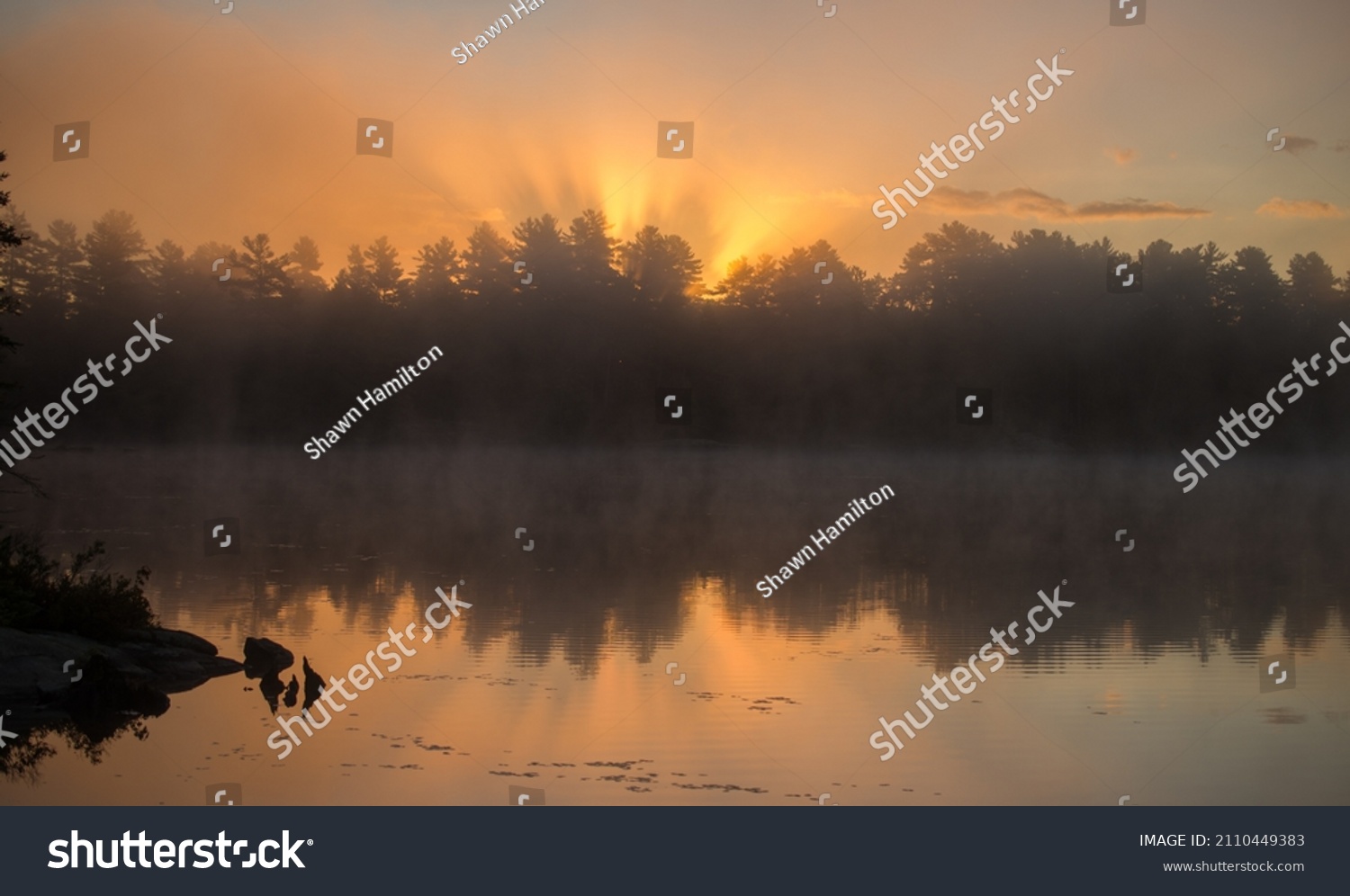 sunrise or sunset behind trees with reflection on calm water of lake at dawn or dusk on lake in cottage country of Ontario horizontal format room for type natural backdrop background or wallpaper  #2110449383
