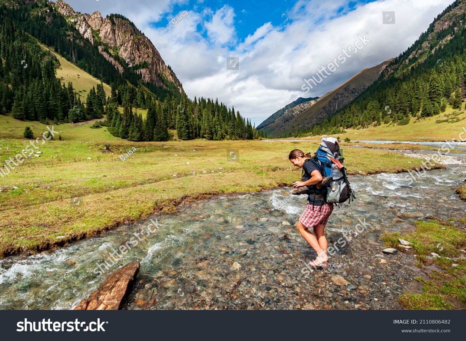 Young woman with big backpack barefoot crossing river. Shoeless female backapcker in river. Karakol valley, Issyk-kul region, Ala-kul lake Terskey Alatau mountain range, Kyrgyzstan, Central Asia. #2110806482