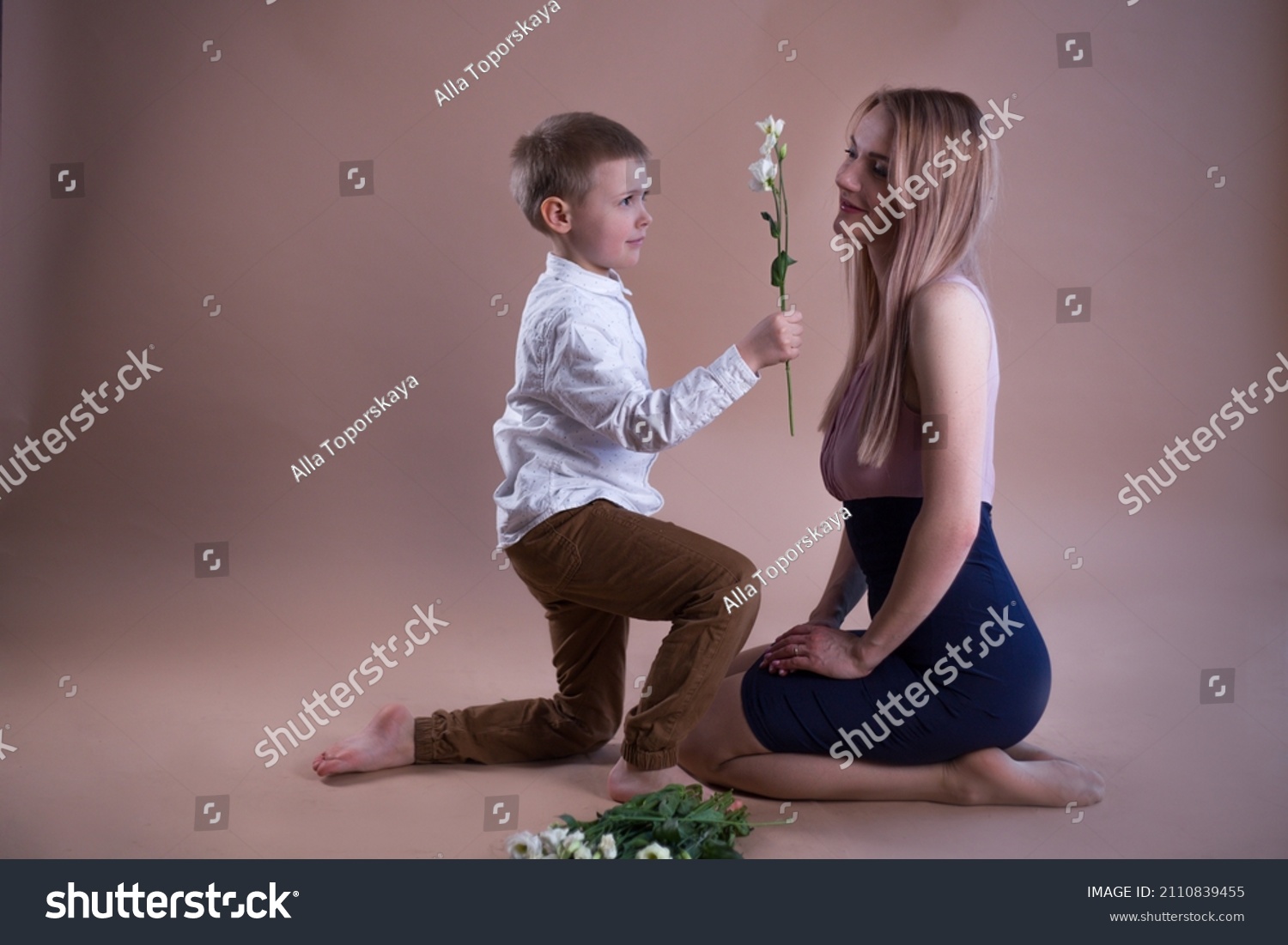 A cute European blonde boy kneels and gives white flowers to a beautiful European girl with blonde hair in the studio on a brown background. #2110839455