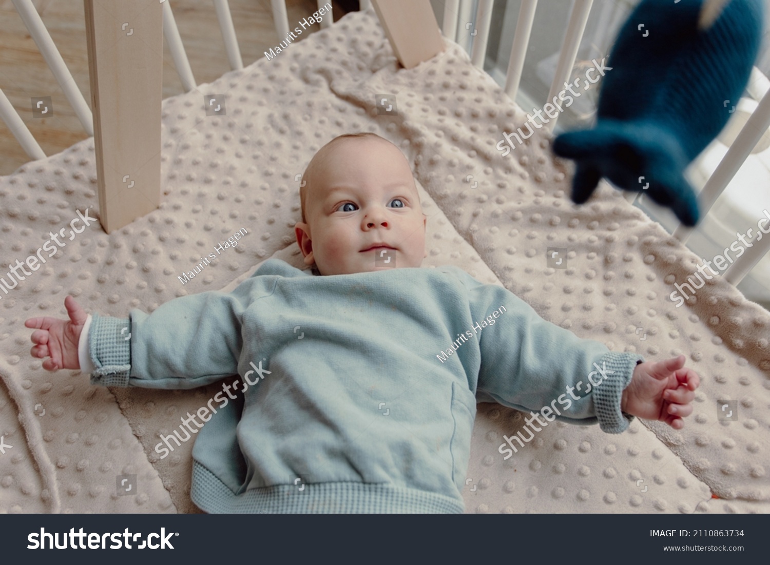 Adorable four month old baby boy laying in play pen looking around smiling. Wears a blue sweater and has blue eyes. #2110863734