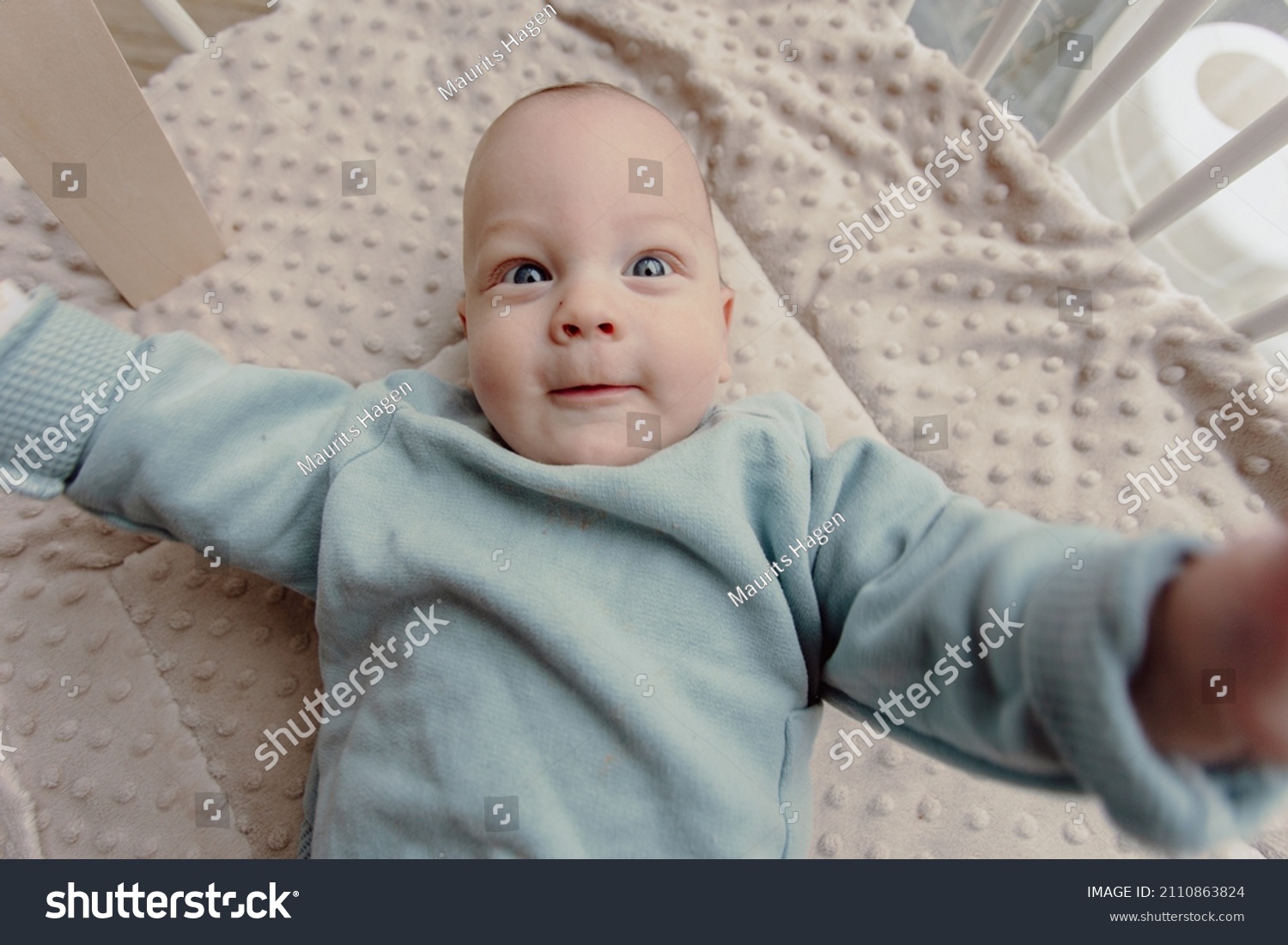 Adorable four month old baby boy laying in play pen looking around smiling. Wears a blue sweater and has blue eyes. #2110863824