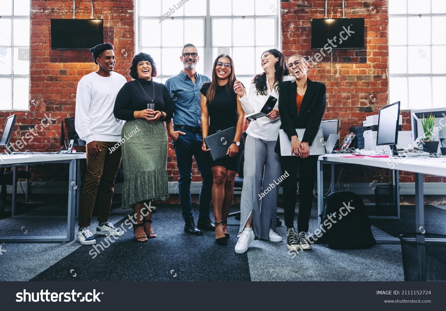 Group of businesspeople laughing cheerfully in a modern workplace. Diverse group of colleagues enjoying working together in an office. Successful businesspeople standing together. #2111152724