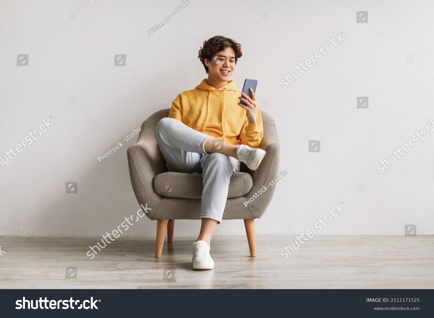 Handsome young Asian guy using cellphone, surfing web or social media, sitting in armchair, enjoying contemporary technologies, checking new mobile app against white studio wall #2111171525