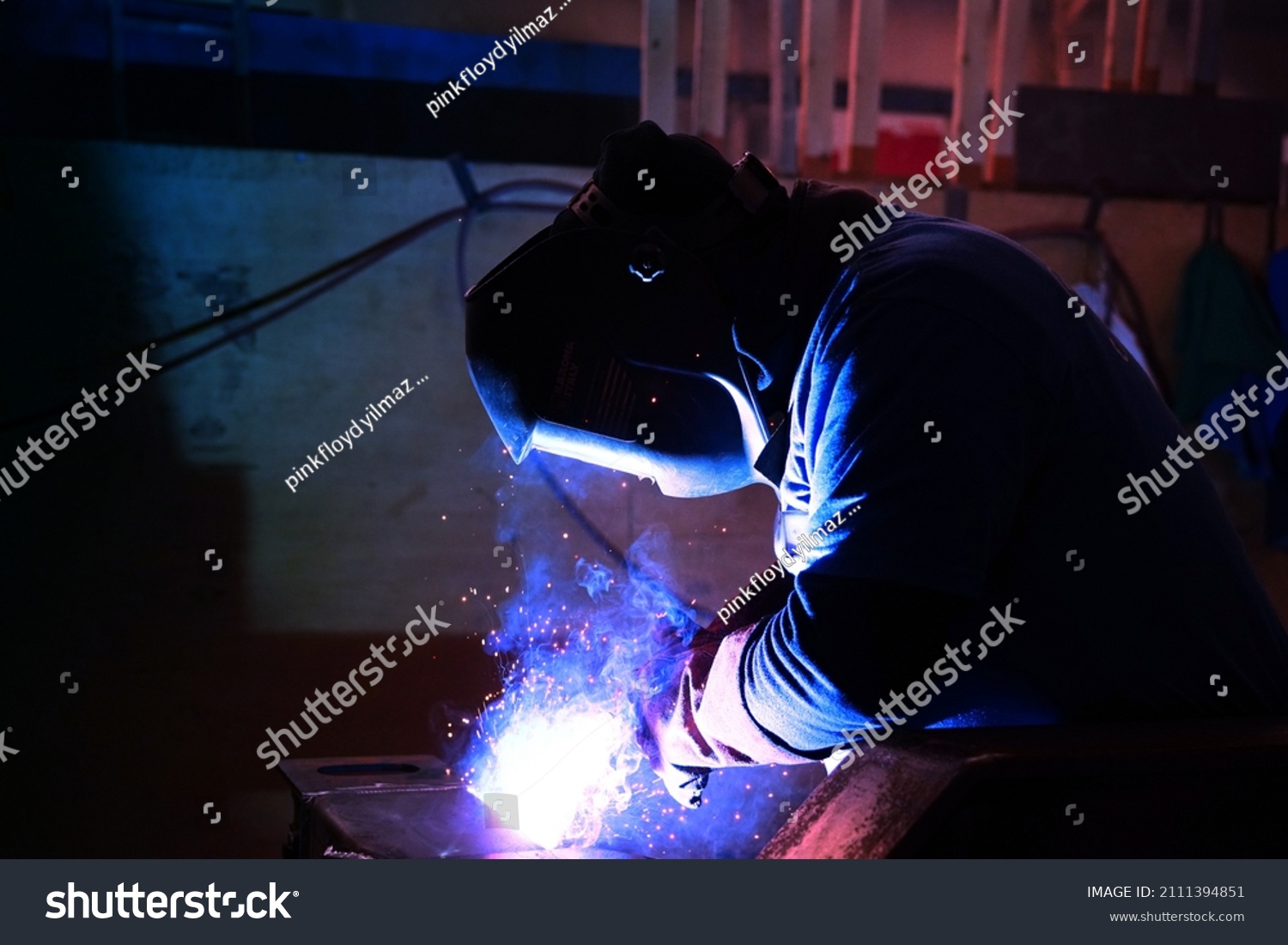 A welder welds metal into his workshop. Blue welding sparks. Gas combustion and blue smoke. Small welding workshop. Welding juncture of metal construction #2111394851