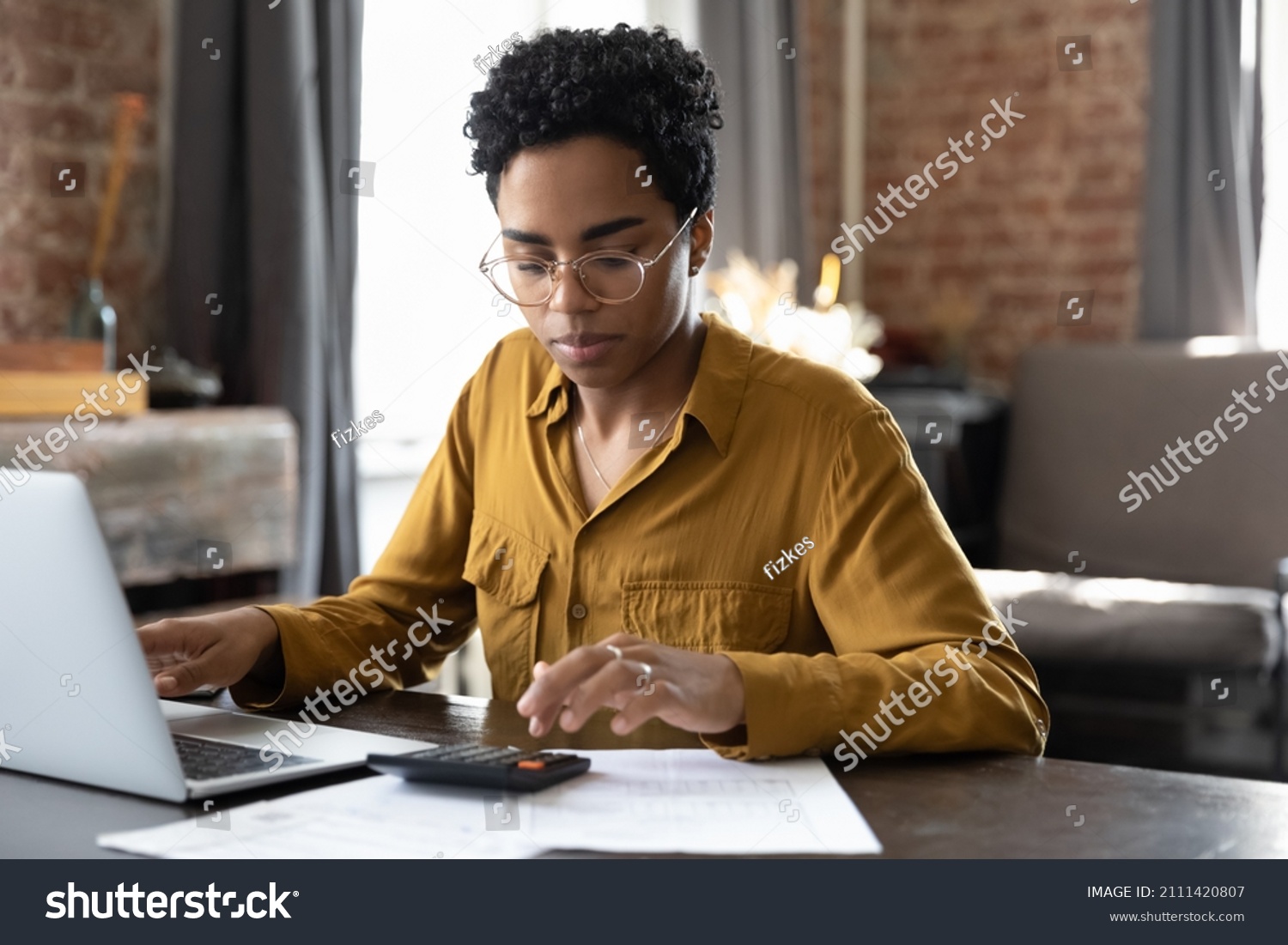 Focused millennial Black business woman calculating finance, money, using calculator, laptop computer at home workplace table, counting budget, paying bills, taxes, rent, mortgage fees #2111420807