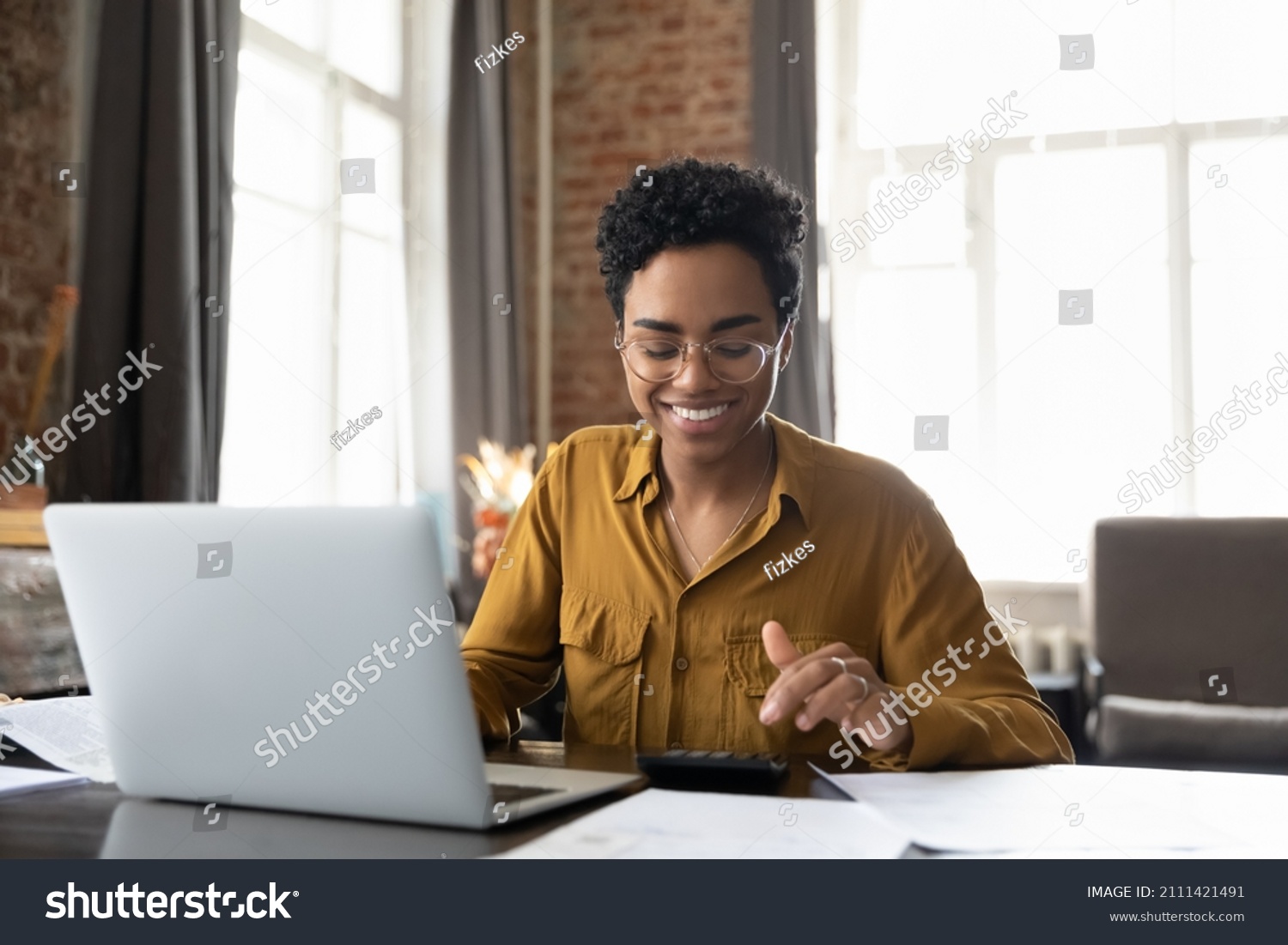 Happy young Afro American entrepreneur woman in glasses counting profit, on calculator at laptop computer, analyzing benefits, enjoying financial success, job high result, smiling #2111421491