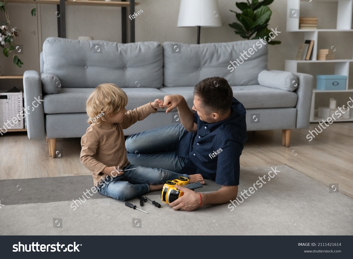 Happy cute small kid son bumping fists with caring millennial father, finishing repairing favorite toy car with screwdriver, having fun improving skills sitting on floor carpet together at home. #2111421614