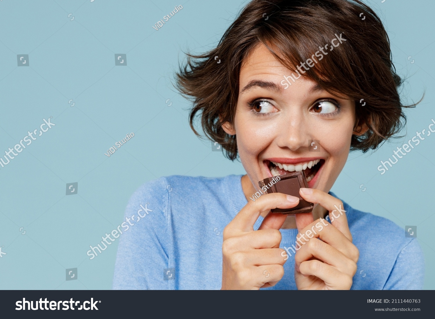 Close up young happy woman in casual sweater hold sweet pink cream donuts biting chocolate bar look camera isolated on plain pastel light blue background studio portrait. People lifestyle food concept #2111440763