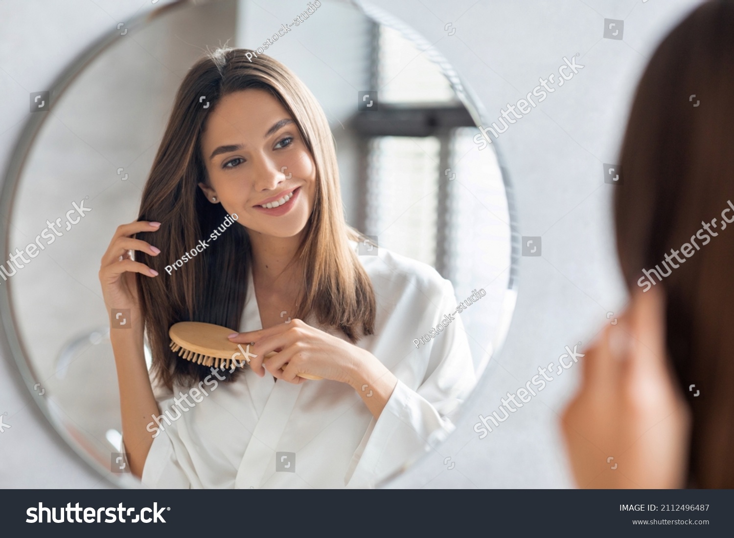 Haircare Concept. Attractive Young Lady Brushing Her Thick Beautiful Hair With Comb While Standing Near Mirror In Bathroom, Happy Woman Wearing White Silk Robe Looking To Her Reflection And Smiling #2112496487