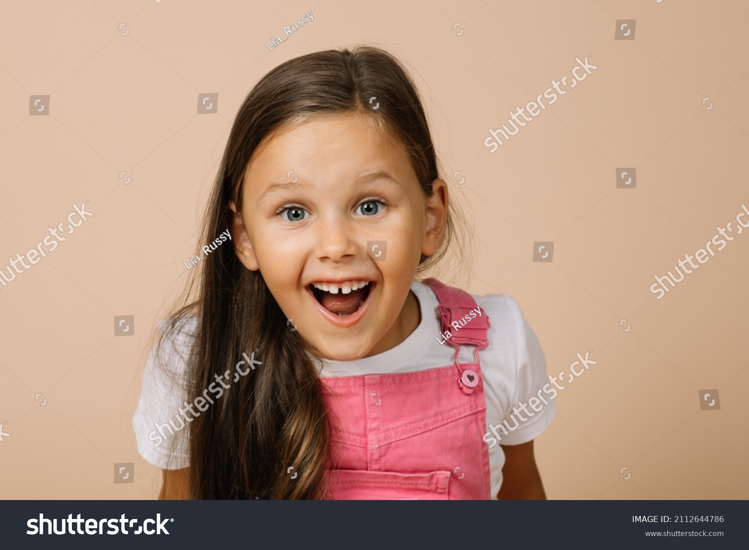 Little girl with surprised eyes, opened mouth, blissful smile with teeth and raised eyebrows looking at camera wearing bright pink jumpsuit and white t-shirt on beige background. #2112644786