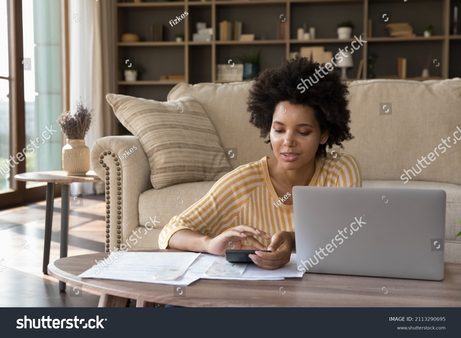 African woman makes calculations on calculator pay utility bills, loan via e-bank application on computer. sit in living room alone. Personal family budget management, modern tech, accounting concept #2113290695