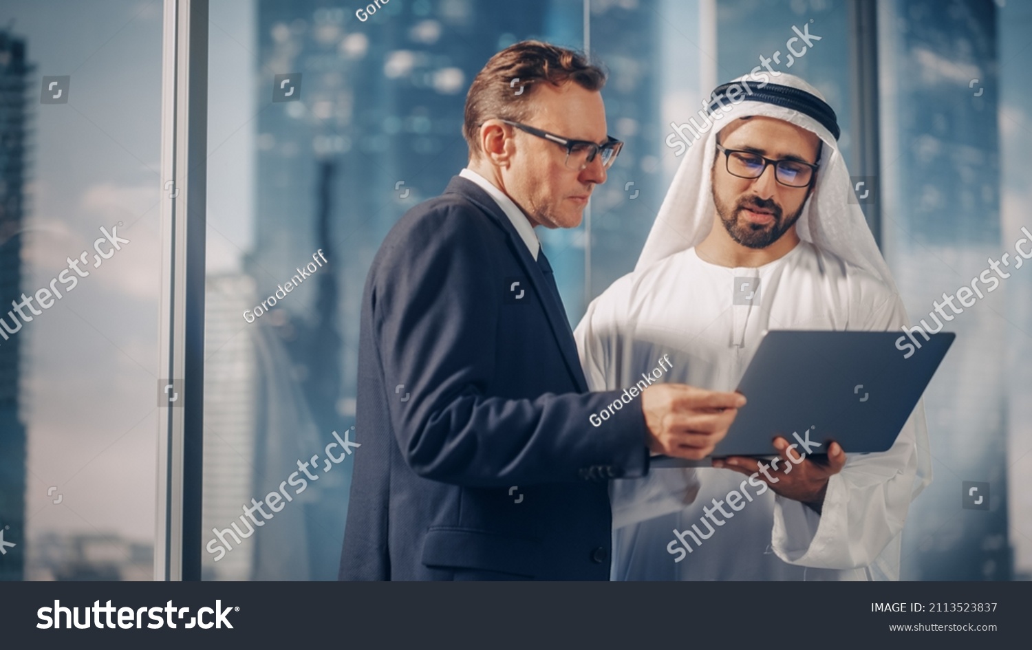 International Operations Manager Meeting Saudi Business Partner in Traditional Kandura. They're Standing in Modern Office, Using Laptop Computer. Successful Saudi, Emirati, Arab Businessman Concept. #2113523837