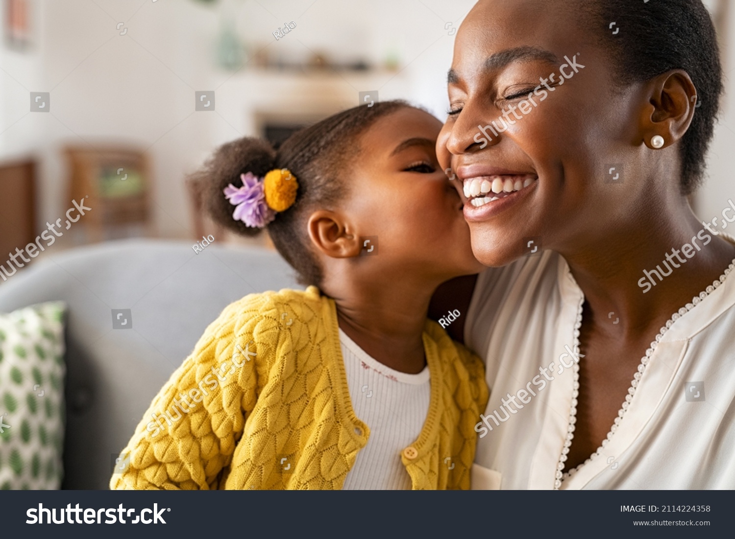 Close up of beautiful daughter kissing mother on cheek at home. African little girl giving kiss to happy mother. Lovely black female child kissing cheerful and proud woman on cheek for mother's day. #2114224358