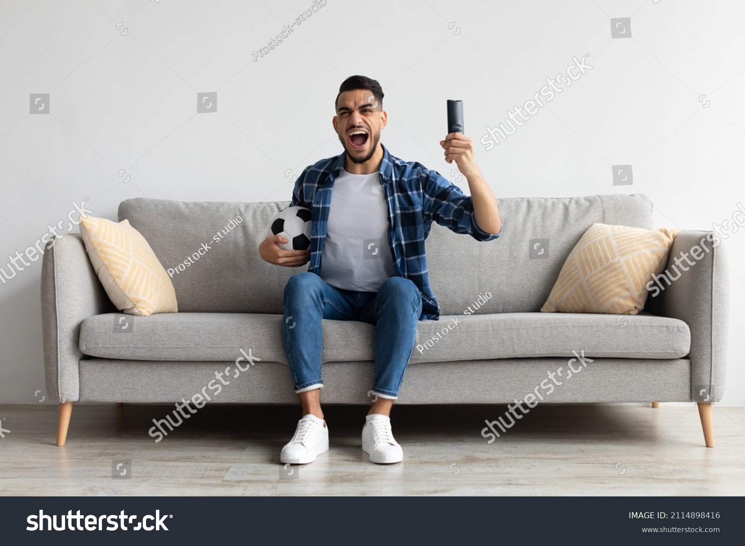 Portrait of excited young guy watching football match, raising clenched fist. Emotional man sitting on couch cheering favorite team enjoying game goal on TV at home holding remote controller and ball #2114898416