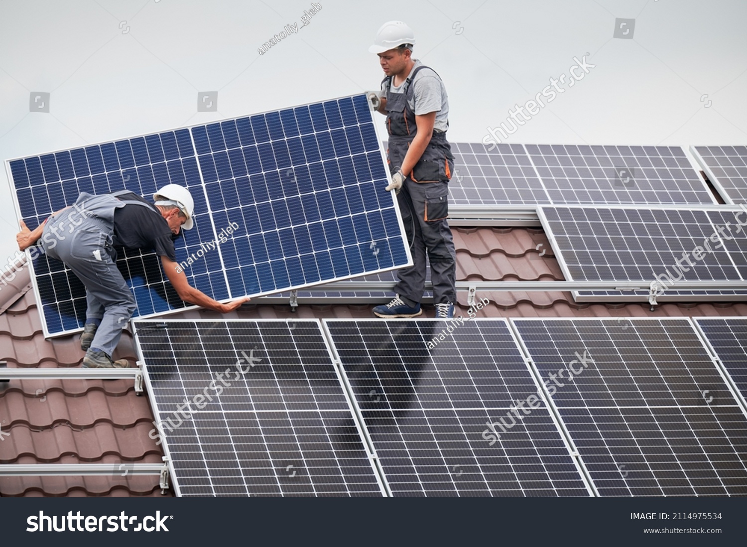 Men technicians carrying photovoltaic solar moduls on roof of house. Engineers in helmets installing solar panel system outdoors. Concept of alternative and renewable energy. #2114975534
