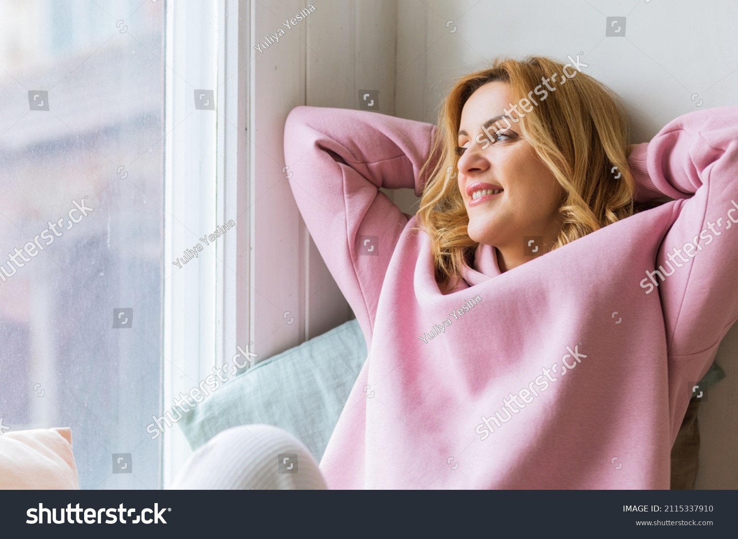 A mature woman in a pink hoodie sits on the windowsill with her arms folded behind her head and enjoys the moment. Slow life concept and digital detox #2115337910