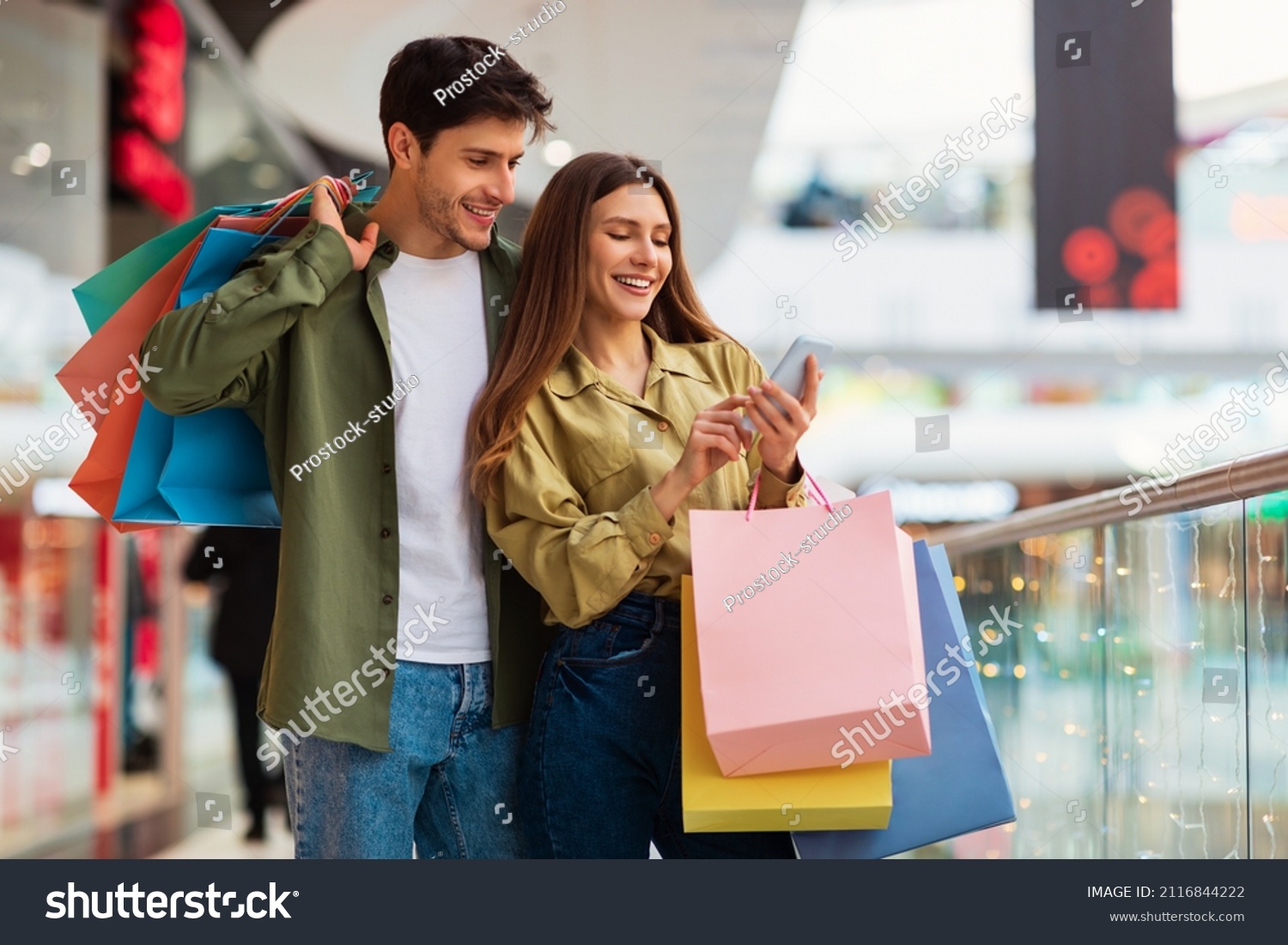 Buyers Couple Shopping Using Cellphone Holding Colorful Shopper Bags Standing In Mall. Happy Customers Using Application Purchasing Clothes Online Via Smartphone. Ecommerce And Shopaholism #2116844222