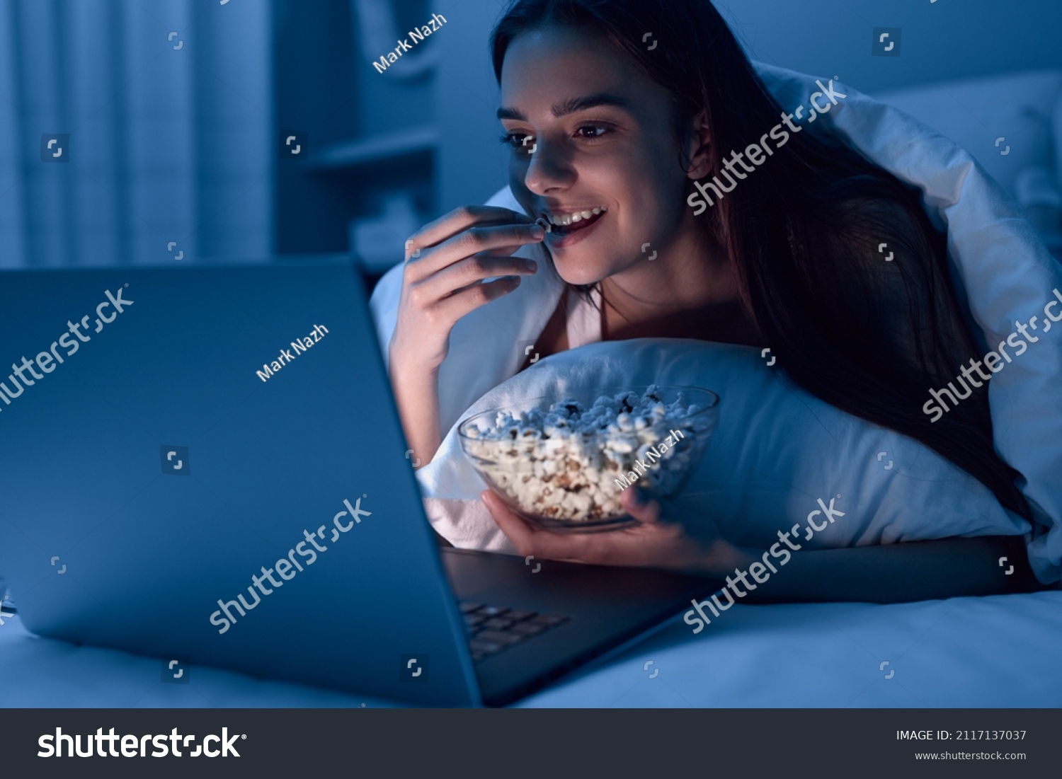 Happy young woman eating popcorn while lying on bed under blanket and watching movie on netbook at night at home #2117137037