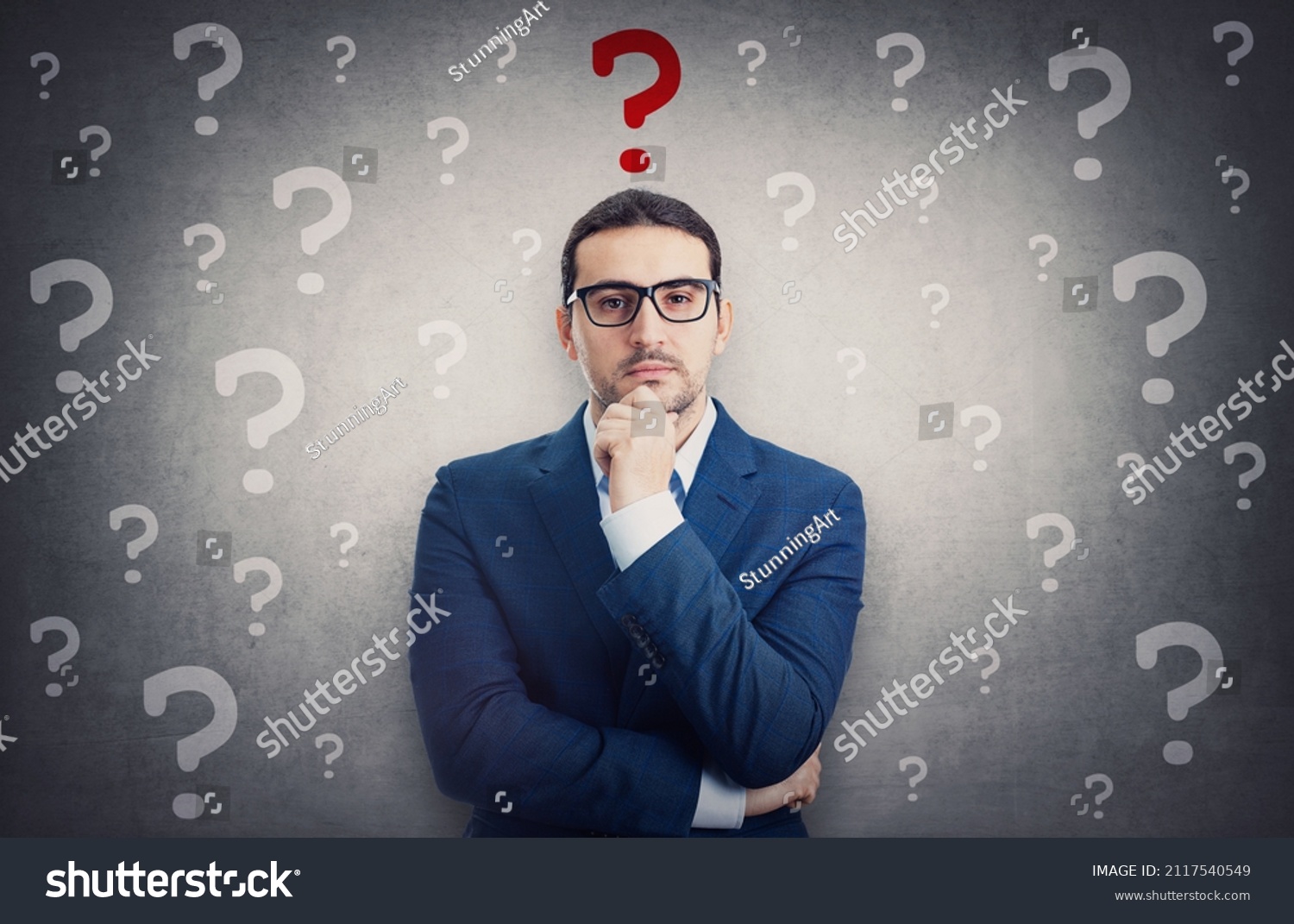 Skeptical businessman surrounded by interrogation marks has a main question as a red symbol over head. Thoughtful business person keeps hand under chin, waiting for an answer #2117540549