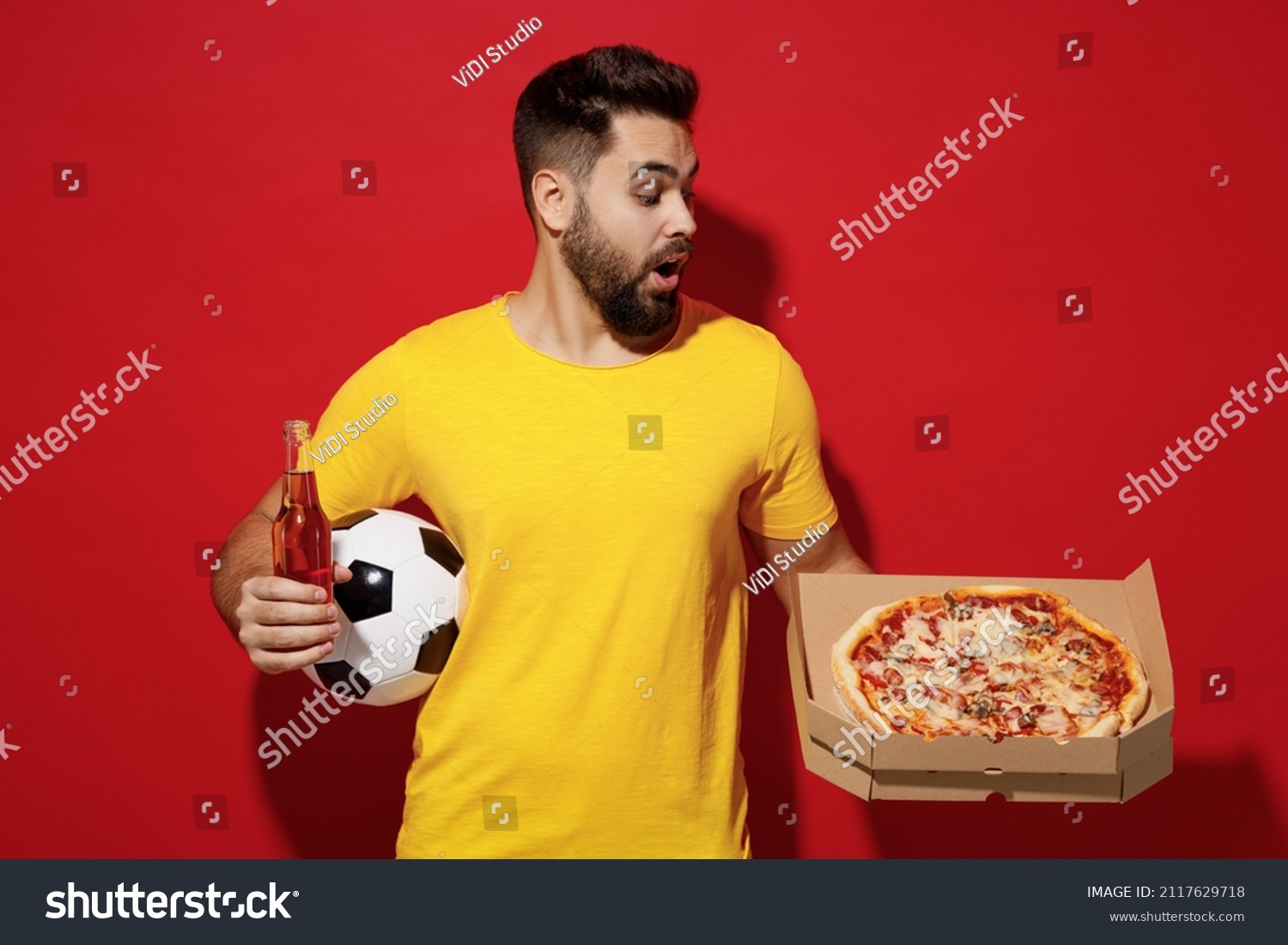 Young bearded man football fan in yellow t-shirt cheer up support favorite team hold soccer ball look at italian pizza in cardboard flatbox beer isolated on plain dark red background studio portrait #2117629718