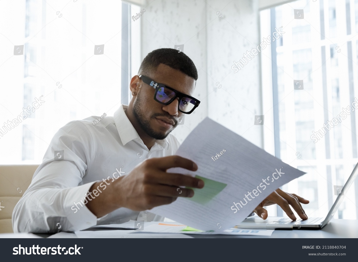 Serious African American manager in stylish glasses reading corporate document with note, sales report, boss order, doing paperwork. Legal expert checking document at workplace with laptop #2118840704