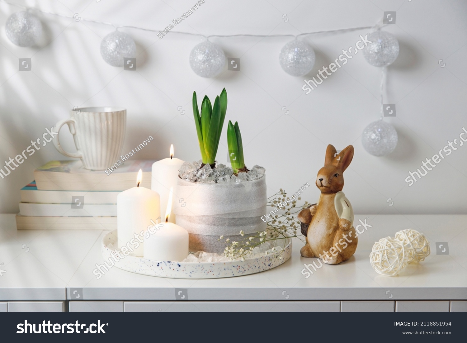 Unblown hyacinths with burning candles on a tray, a stack of books, a mug of tea, a garland of thread on the wall. Easter room decoration. #2118851954
