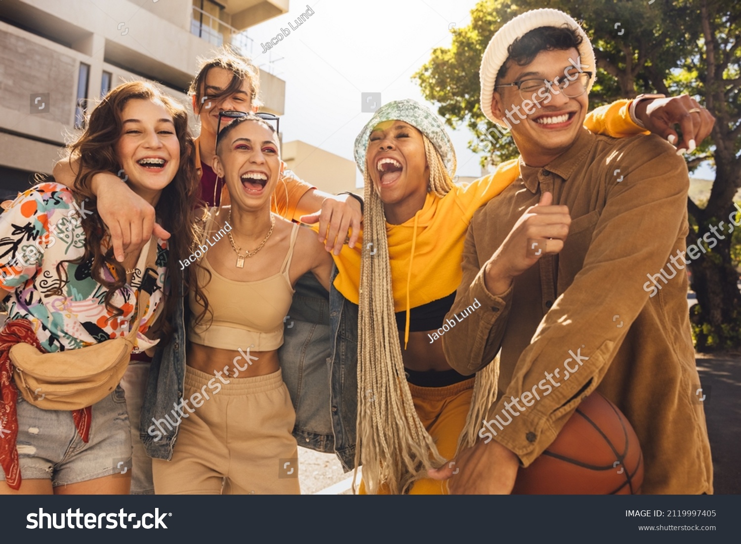 Group of generation z friends laughing together outdoors. Cheerful young friends embracing each other in the summer sun. Youngsters having fun and enjoying their youth. #2119997405