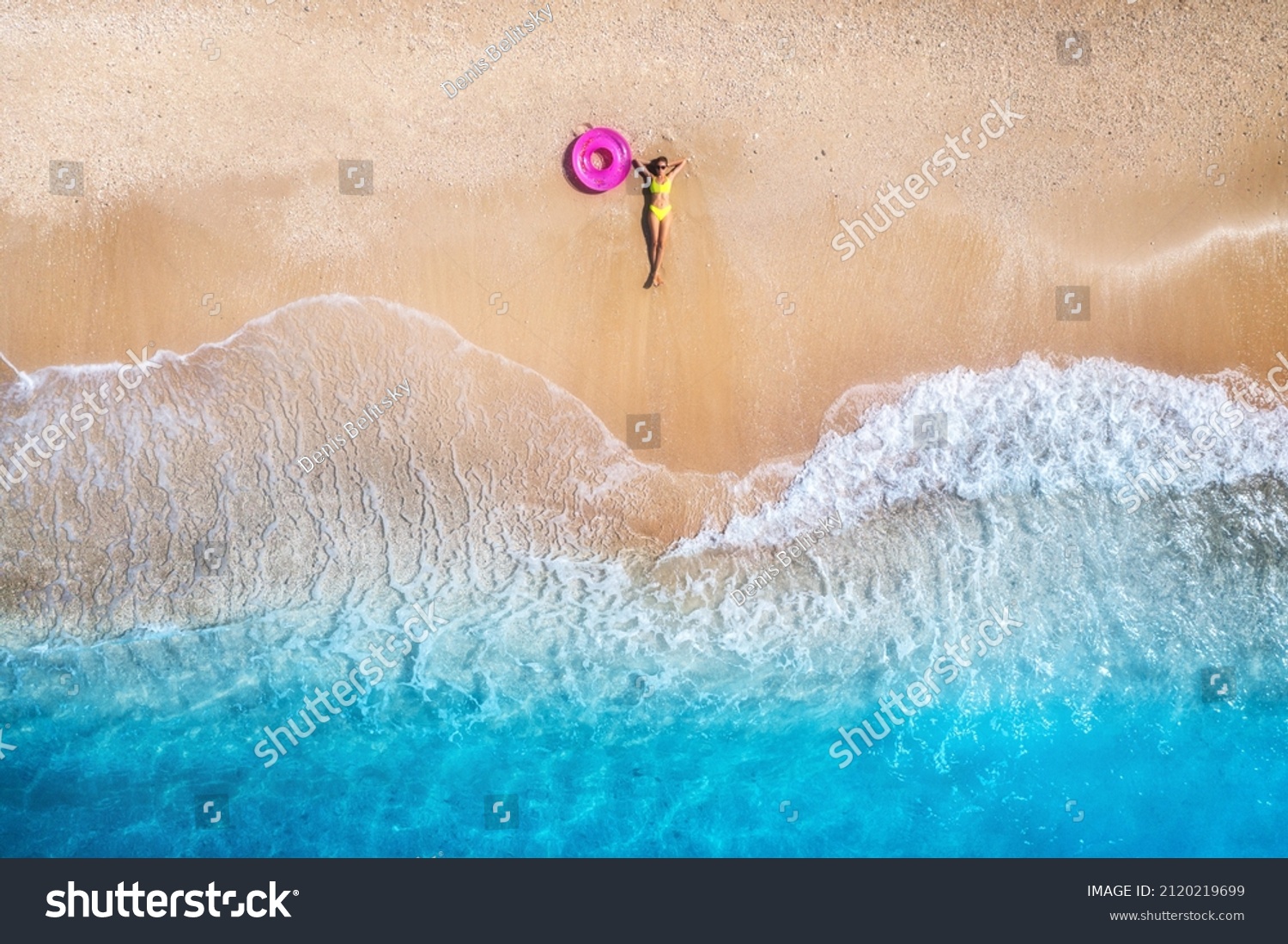 Aerial view of the lying beautiful young woman with pink swim ring on the sandy beach near sea with waves at sunset. Summer vacation in Lefkada island, Greece. Top view of slim girl, clear azure water #2120219699