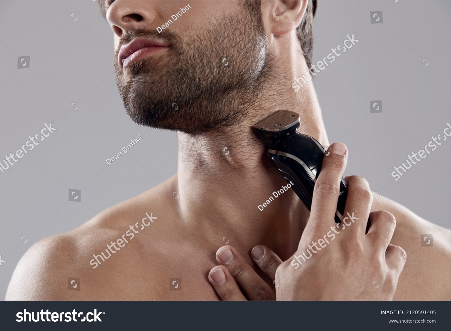 Obscure face of young man shaving beard with electric razor. Handsome dark-haired bearded guy. Concept of face care. Idea of male beauty. Isolated on white background. Studio shoot #2120591405