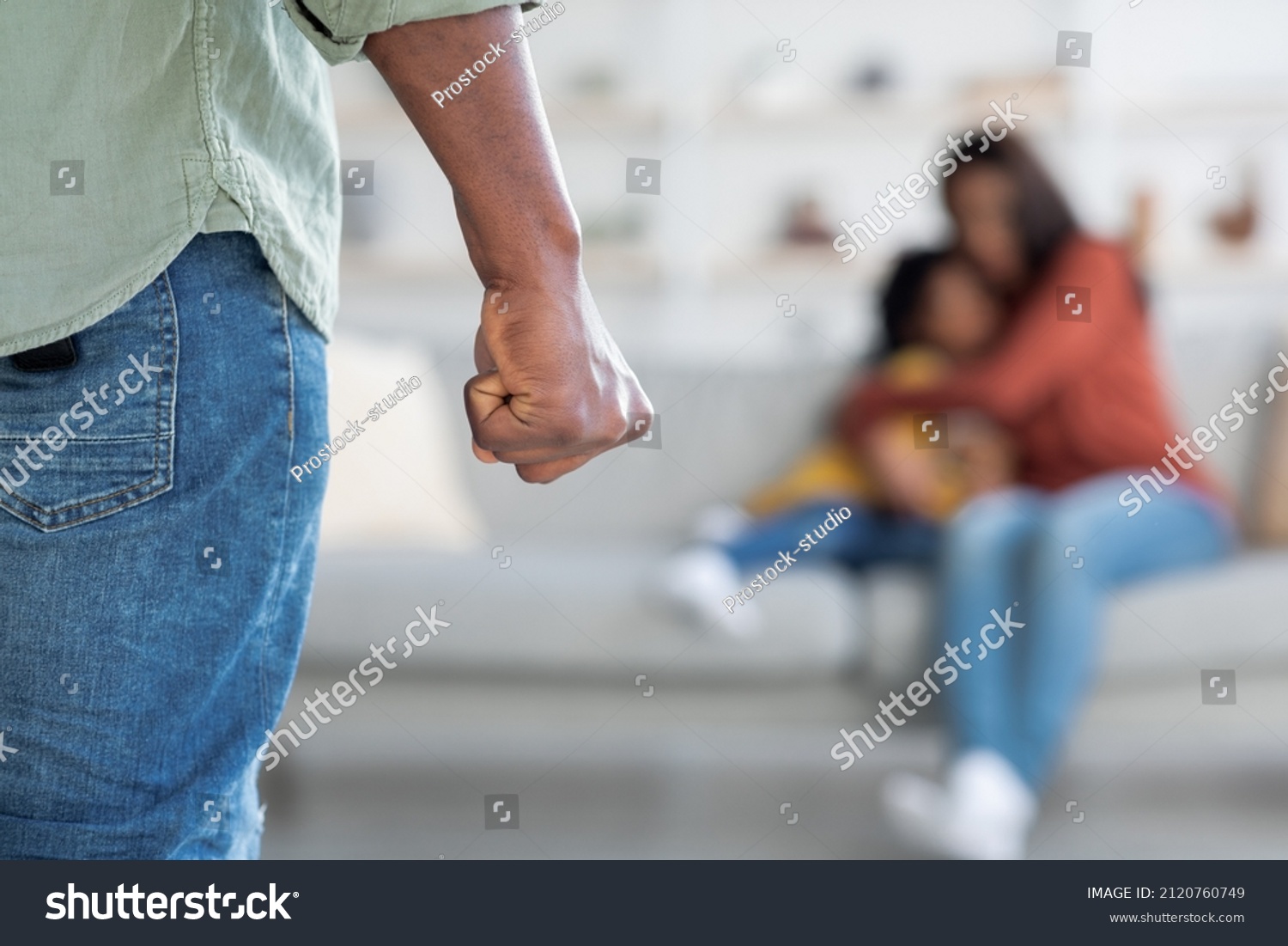 Domestic Violence. Unrecognizable African American Man Threatening Wife And Daughter With His Fist, Scared Mother Embracing Little Girl While Sitting Together On Couch, Selective Focus On Male Hand #2120760749