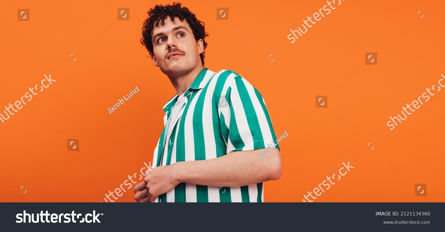 Young queer man looking away thoughtfully in a studio. Confident young generation z hipster standing alone against an orange background. #2121134360