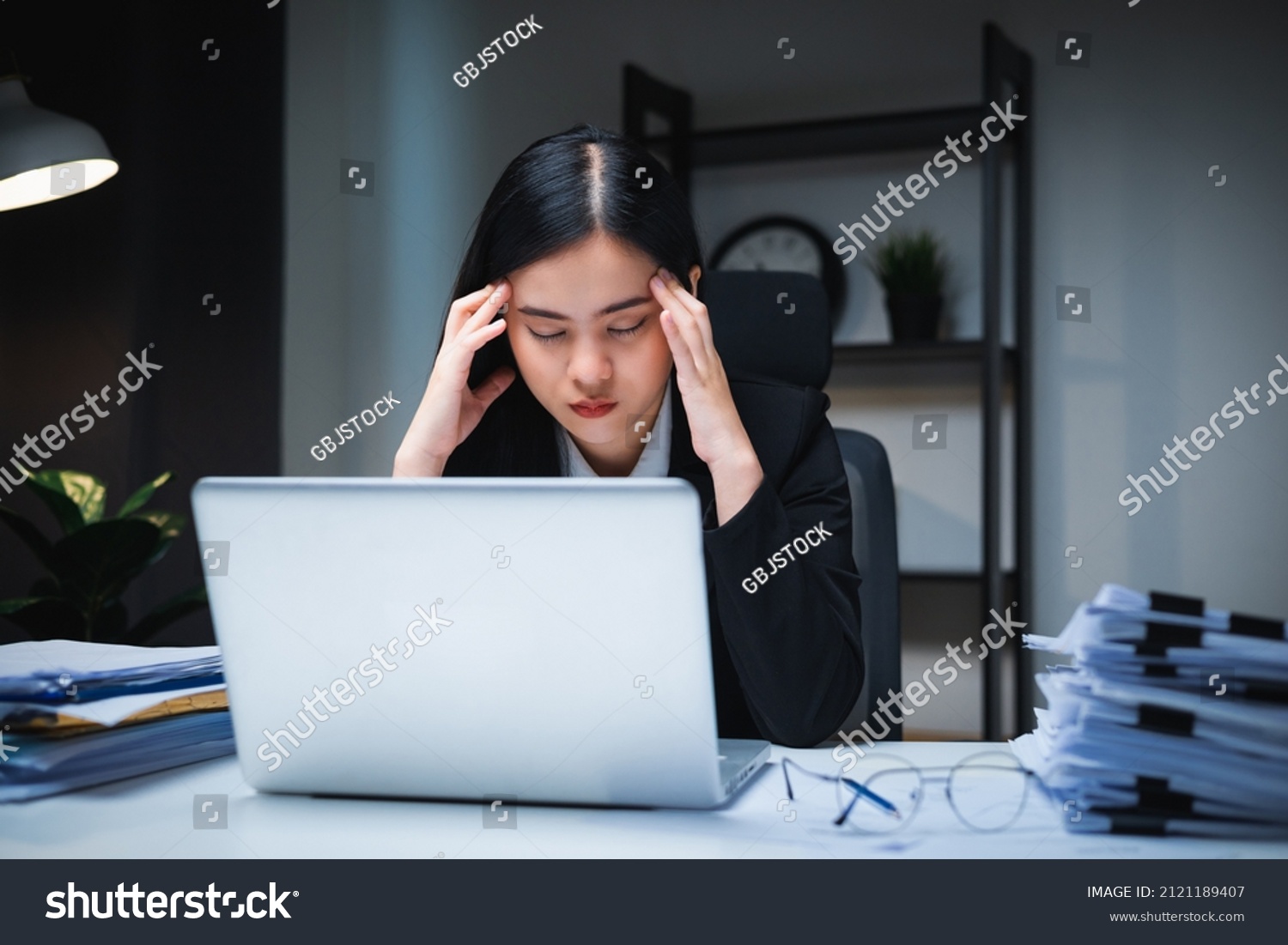 Stressed asian business woman working late at night in the office hands on head feeling headache. Tired woman looking at laptop working hard sitting in the dark room office. Overtime concept #2121189407