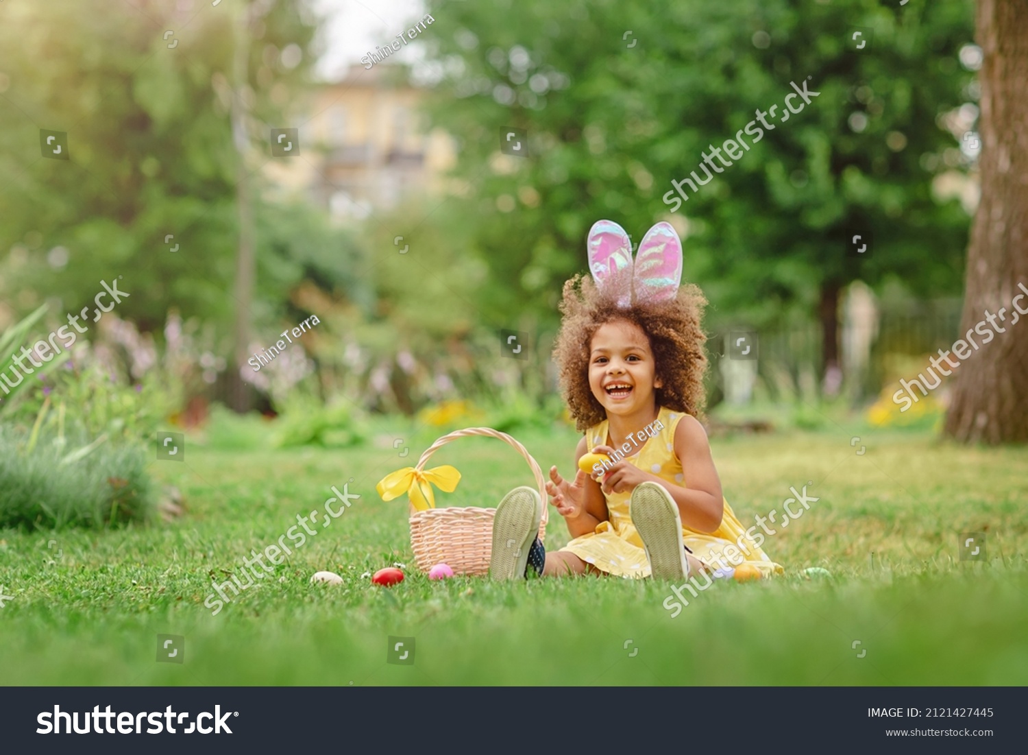 Little Black girl wear bunny ears and gathering Easter eggs on Easter egg hunt in garden #2121427445