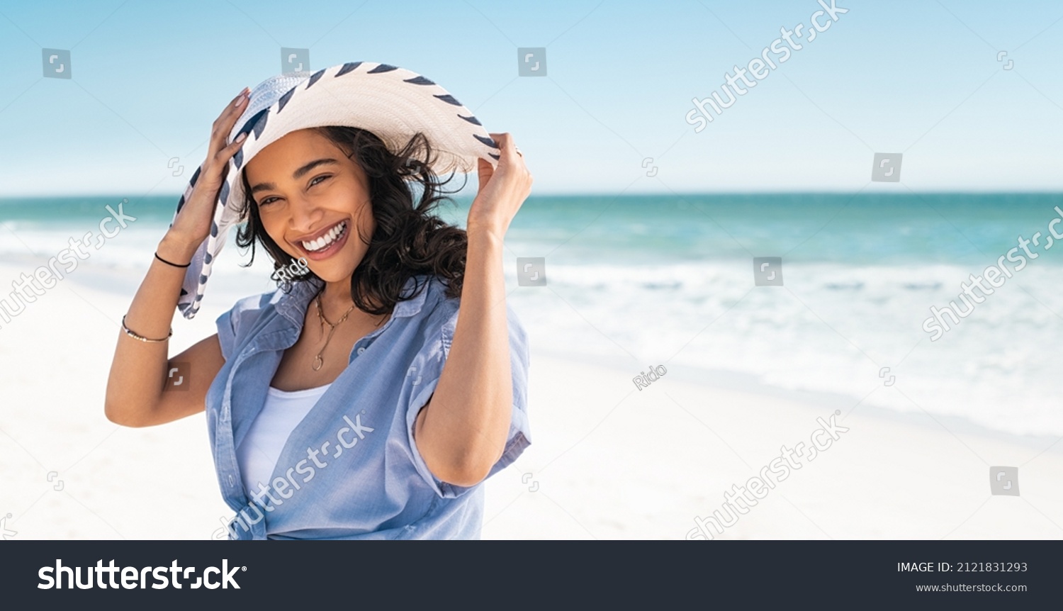 Portrait of stylish latin hispanic woman with white straw hat standing at beach. Young smiling woman on vacation enjoy sea breeze wearing straw hat and looking at camera. Attractive beautiful girl. #2121831293
