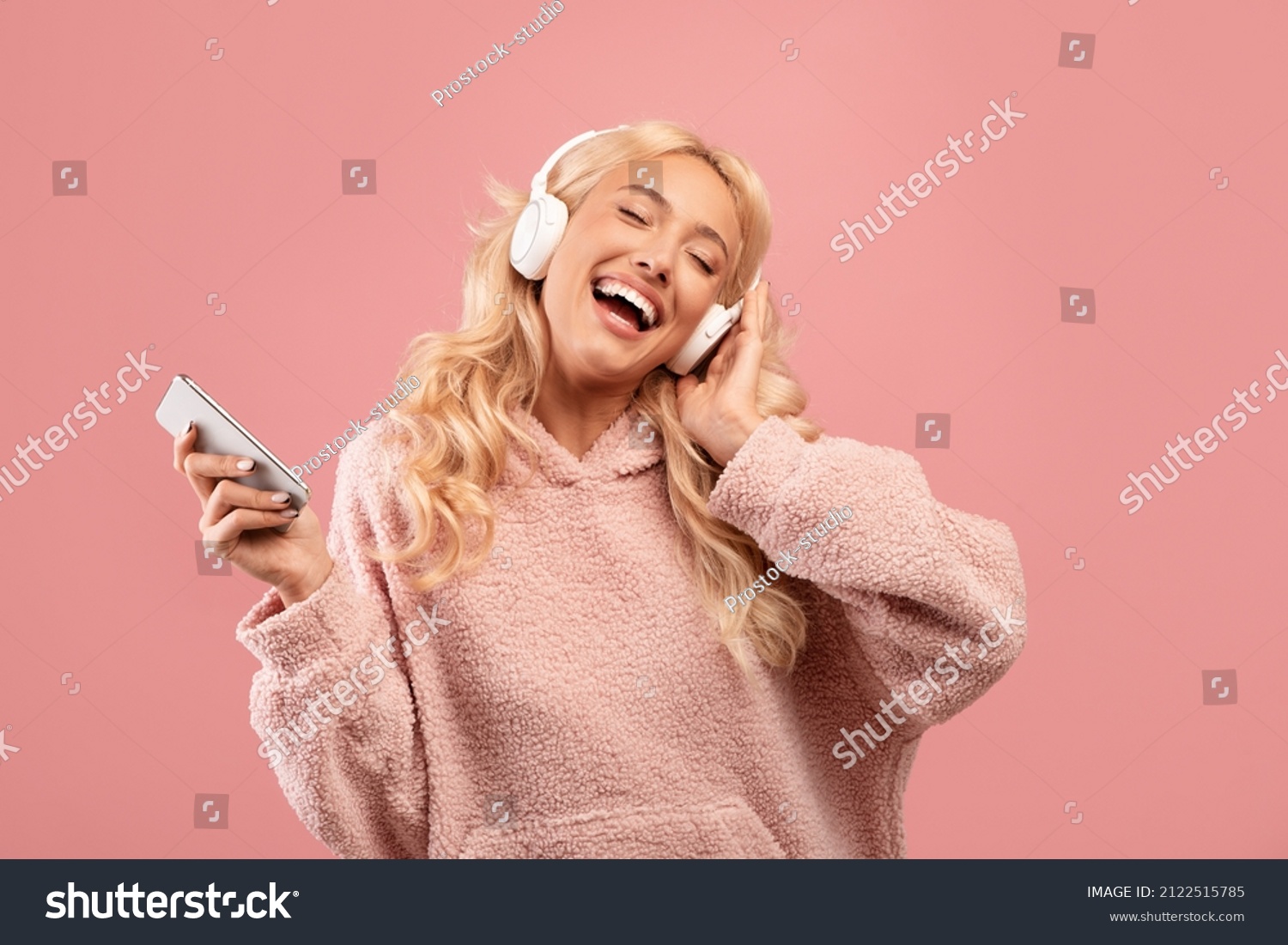 Overjoyed happy lady with headphones and smartphone listening to music and dancing, enjoying cool playlist and moving to favorite song over pink background #2122515785