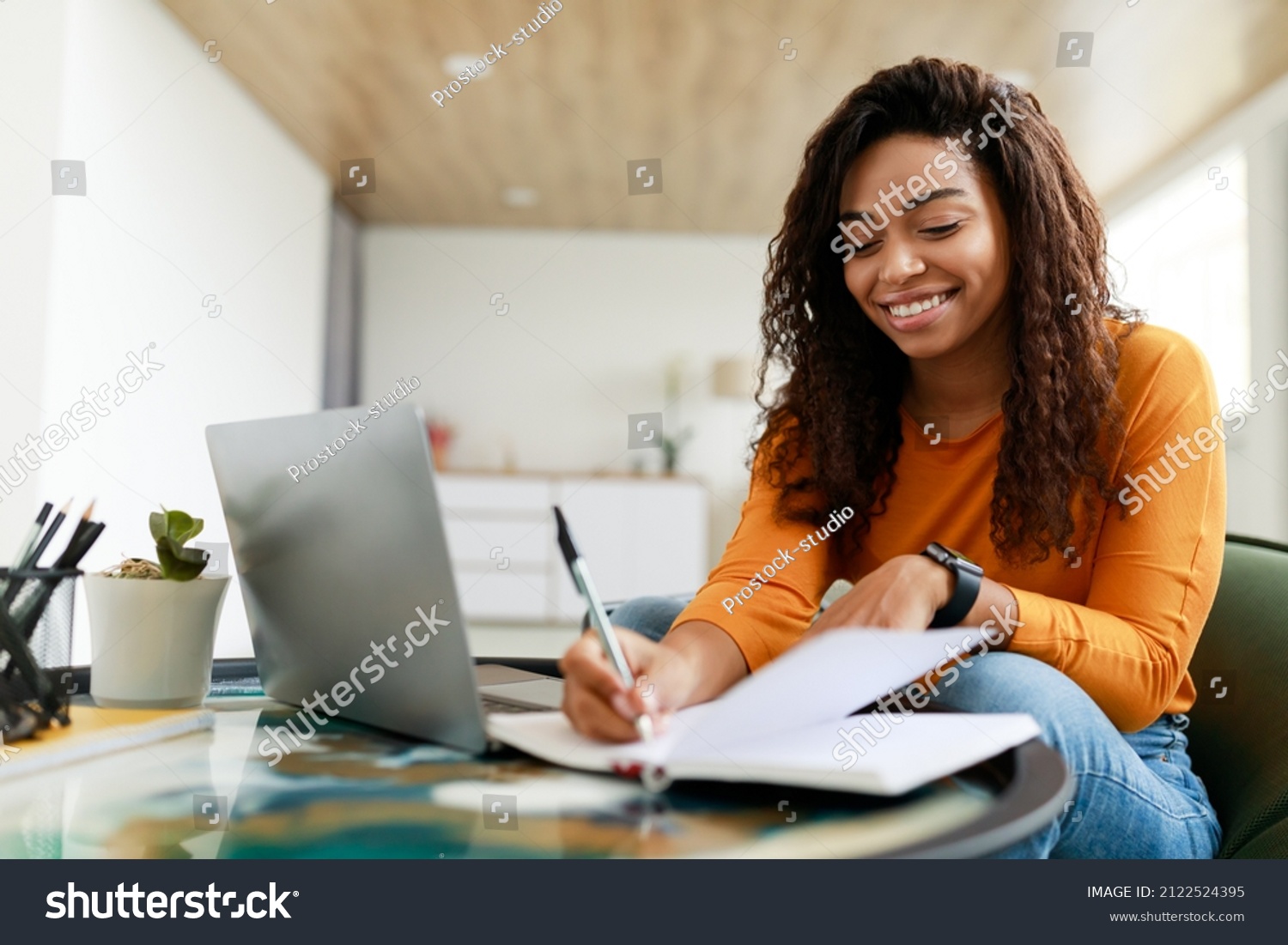 Business And Education Concept. Smiling young black woman sitting at desk working on laptop writing letter in paper notebook, free copy space. Happy millennial female studying using pc #2122524395
