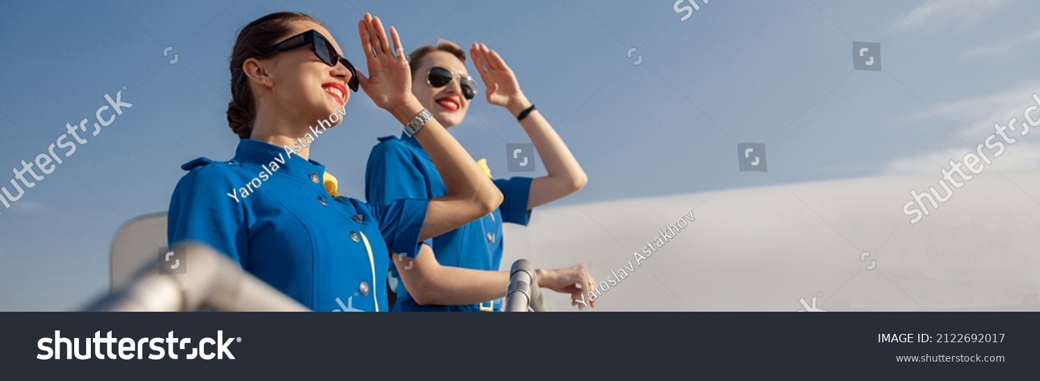 Portrait of two elegant air stewardesses in blue uniform and sunglasses covering eyes with hand and looking far away, standing together on airstair. Aircrew, occupation concept #2122692017