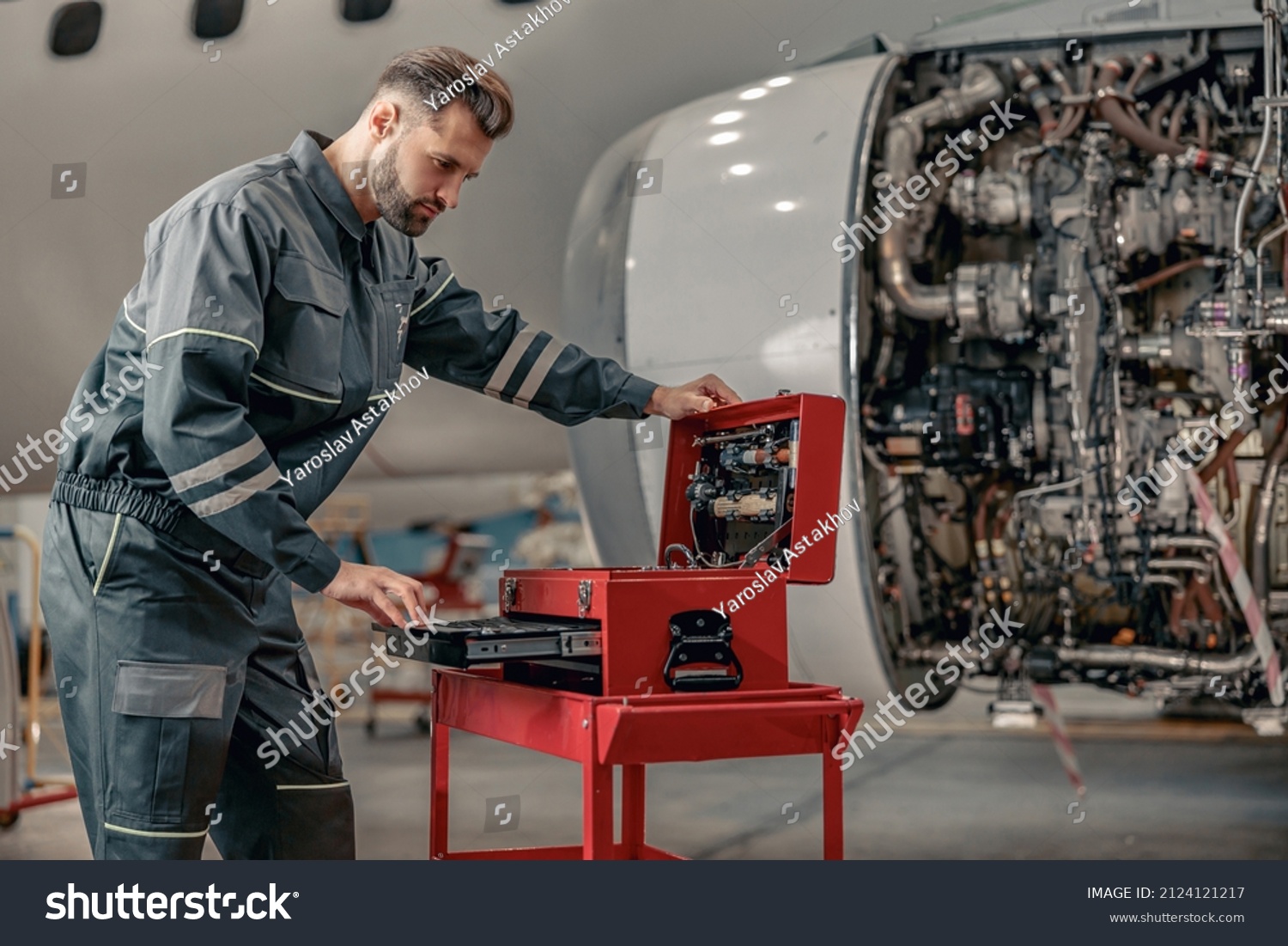 Bearded man airplane maintenance technician in work uniform checking box with instruments while repairing plane #2124121217