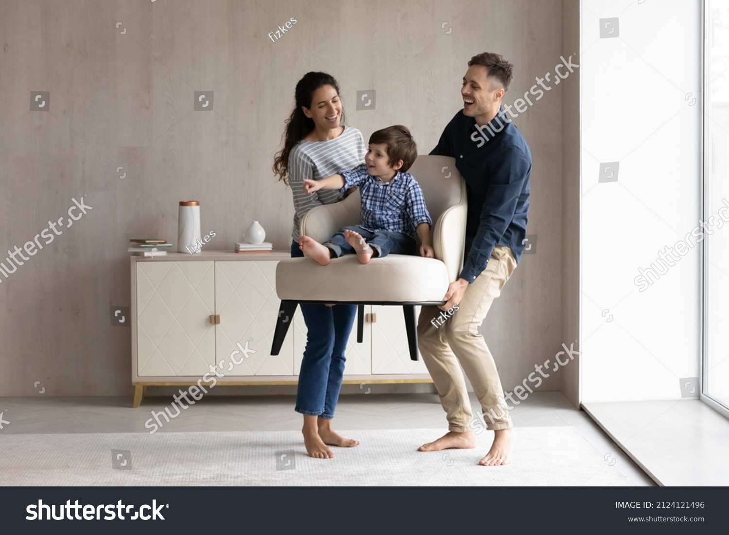 Couple of happy funny parents holding cheerful little son sat in armchair. Family moving into new house, carrying furniture, feeling joy. Mom and dad playing active games with kid. Relocation concept #2124121496